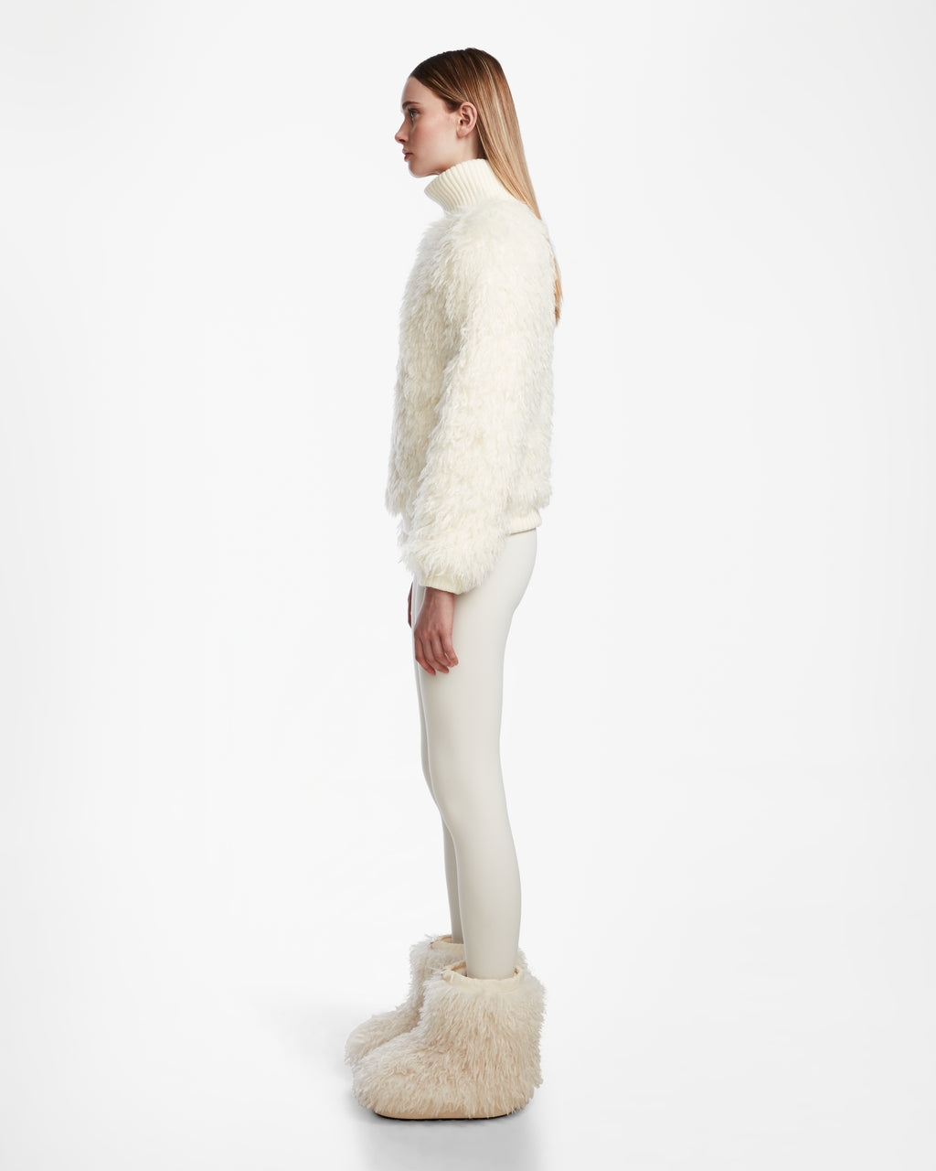 A woman in profile against a white background wears the Rudsak LAYLA M - 8524600 Iceland jacket, styled with fitted white pants and oversized furry white boots for a chic après-ski look.