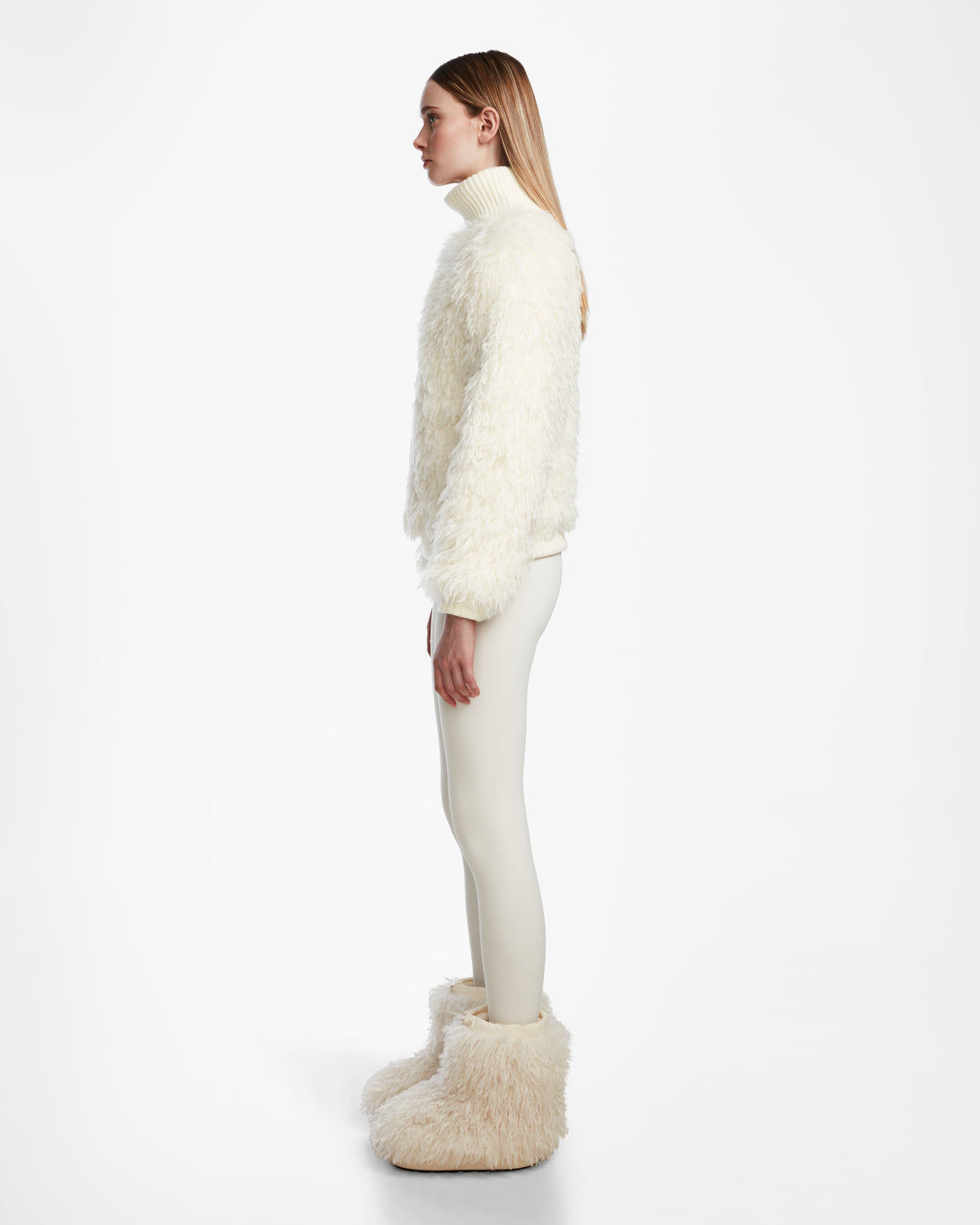 A woman in profile against a white background wears the Rudsak LAYLA M - 8524600 Iceland jacket, styled with fitted white pants and oversized furry white boots for a chic après-ski look.