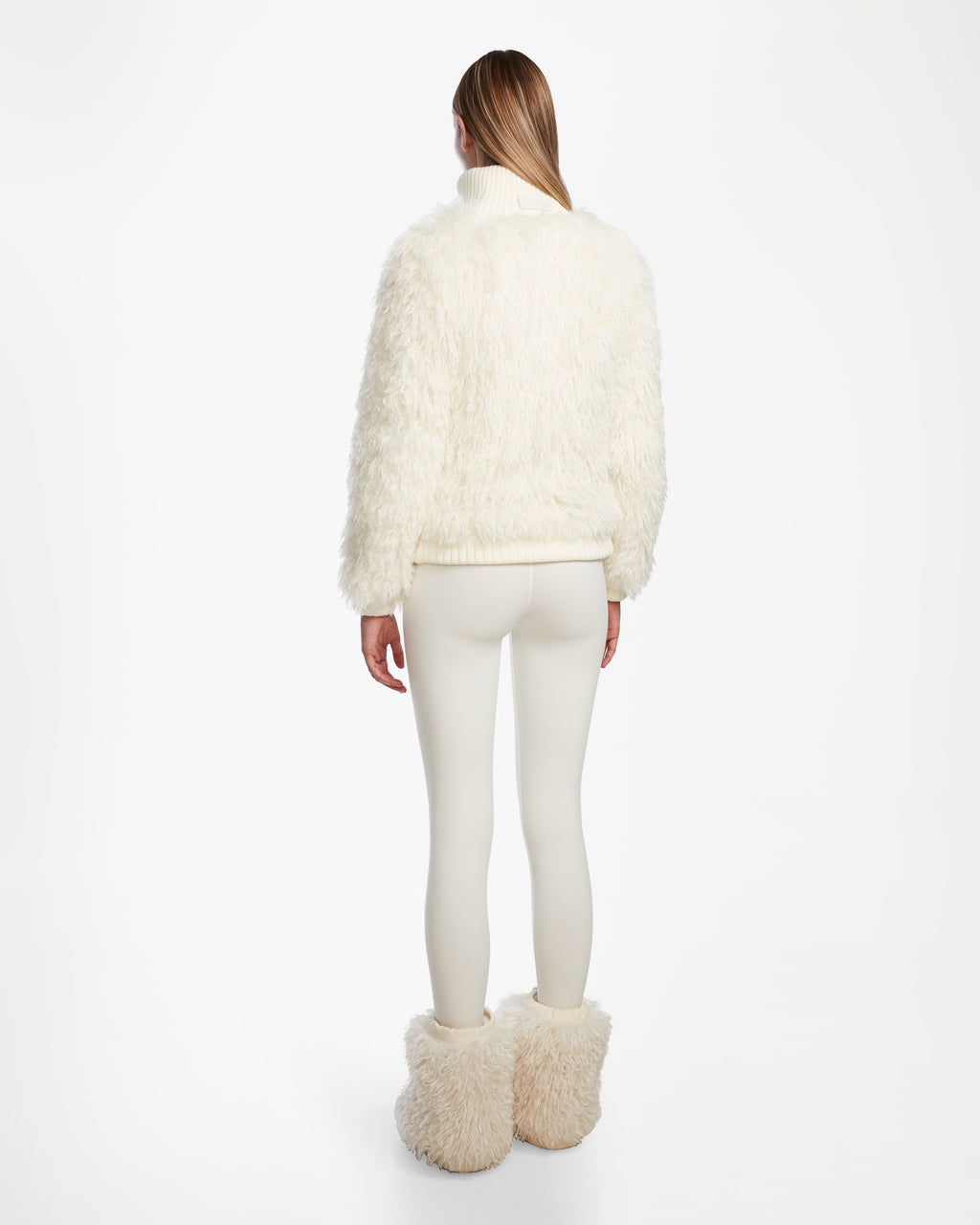 A woman faces away, dressed in Rudsak’s LAYLA M - 8524600 Iceland faux Mongolian fur turtleneck, matching fluffy boots, and cream leggings—an elegant après-ski look set against a plain white background.