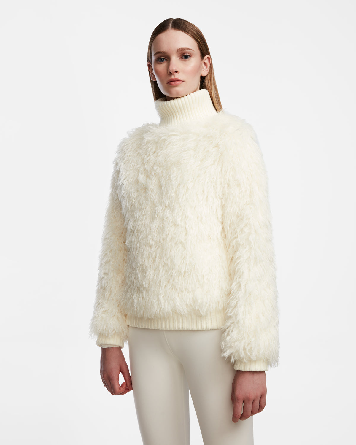 A woman poses against a white background in the Rudsak LAYLA M - 8524600 Iceland après-ski look, wearing a cream faux Mongolian fur turtleneck sweater and matching fitted pants, gazing directly at the camera with a neutral expression.