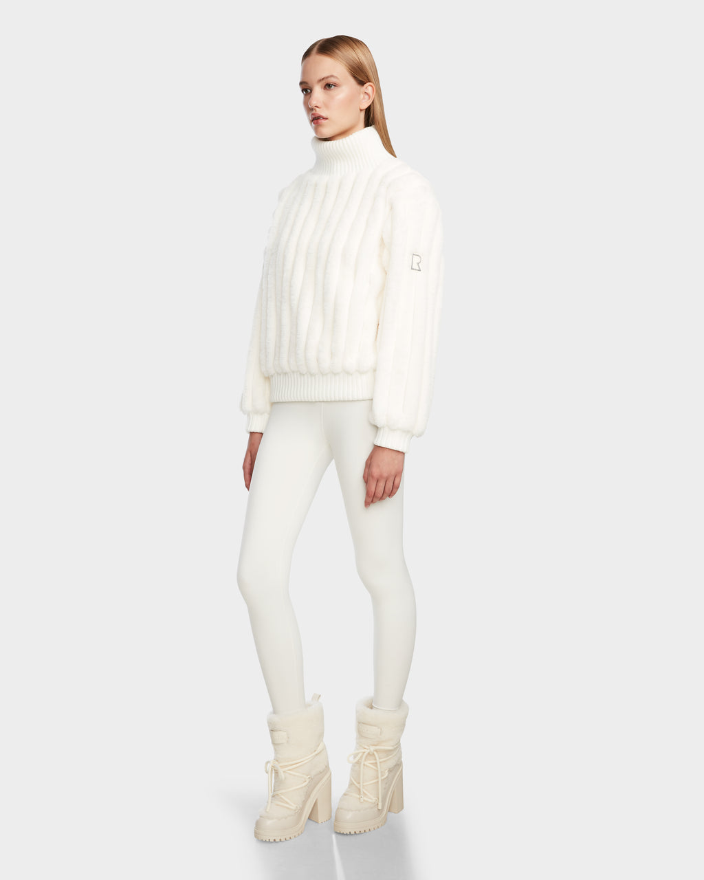 A woman poses against a light gray background in the LAYLA - 8524610 Iceland turtleneck by Rudsak, styled with white leggings and cream winter boots. Her straight blonde hair and neutral expression complete the chic look.
