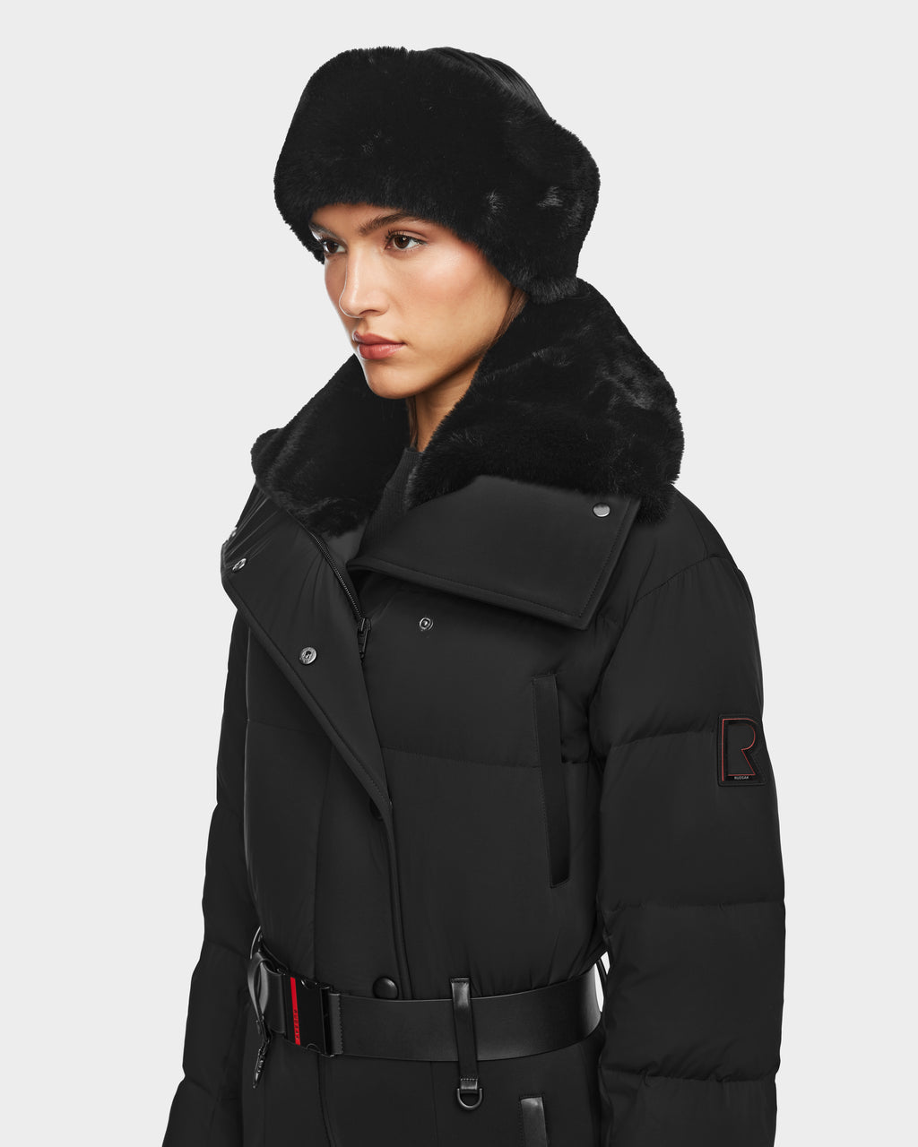A woman wears the Rudsak LOVE - 8124724 Black puffer coat with a wide fur collar and belted waist over a sleek ski suit, looking to the side against a plain white background.