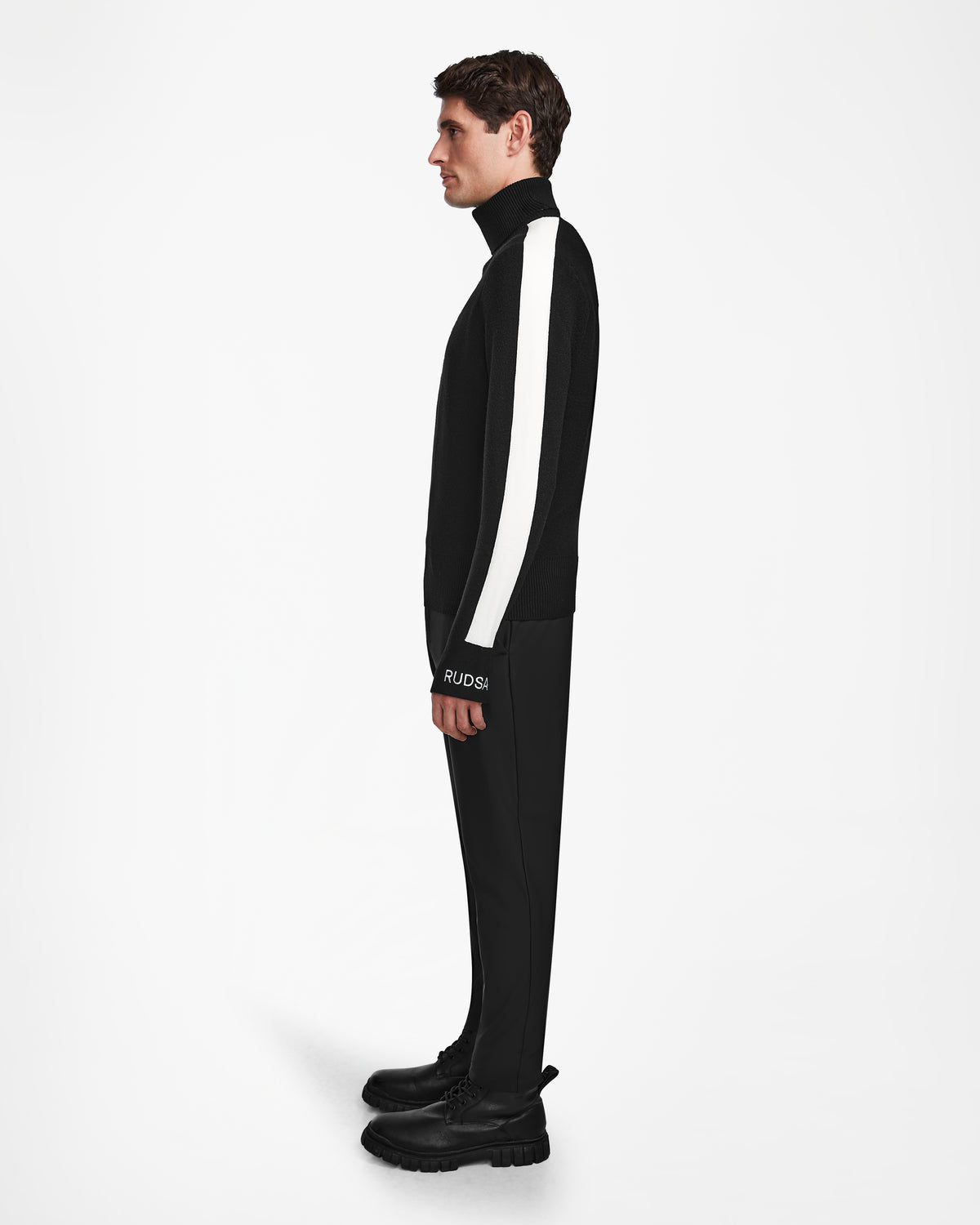 A man in profile against a white background wears the Rudsak LUKAS - 6524543 Black mens knit turtleneck sweater with a bold white sleeve stripe—ideal for après-ski or winter sports-inspired looks.