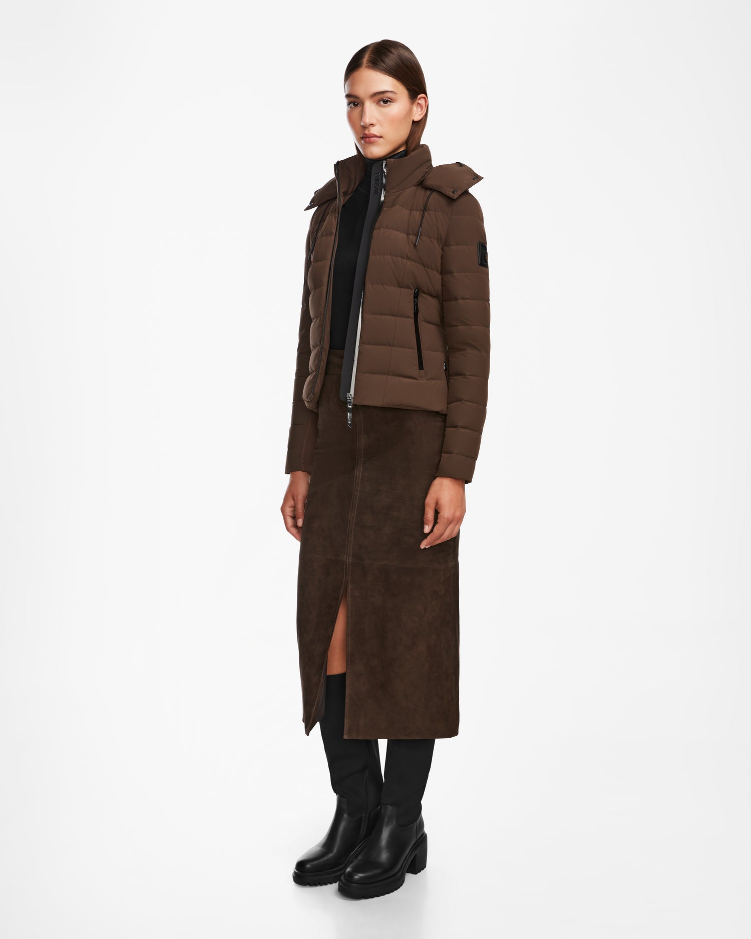 A woman with straight brown hair and a neutral expression stands against a white background wearing the Rudsak MELISSA - 8124824 Espresso down puffer jacket, a long brown suede skirt with front slit, black top, and black boots.