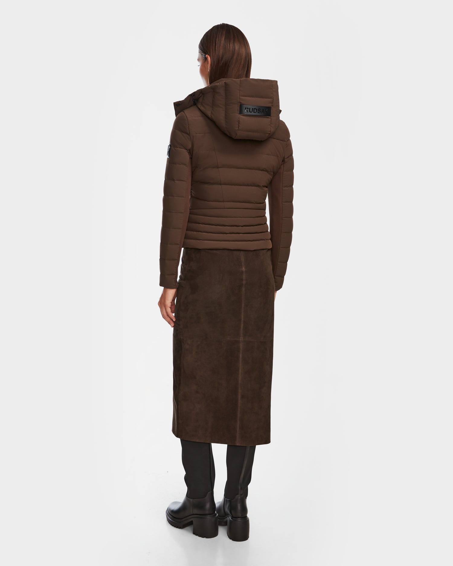A woman stands with her back to the camera in the Rudsak MELISSA - 8124824 Espresso lightweight down puffer jacket, paired with a brown suede midi skirt and black knee-high boots against a plain white background.