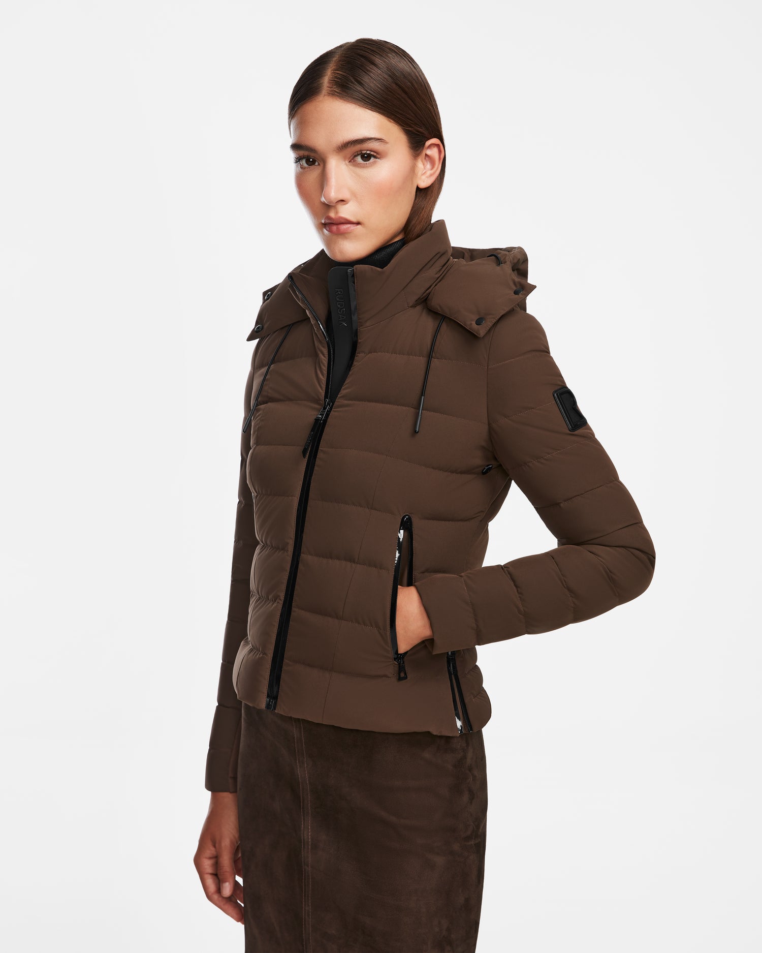 A woman with straight brown hair wears the Rudsak MELISSA - 8124824 Espresso fitted, weatherproof down jacket with hood and zippered pockets, paired with a brown skirt, standing against a plain white background.