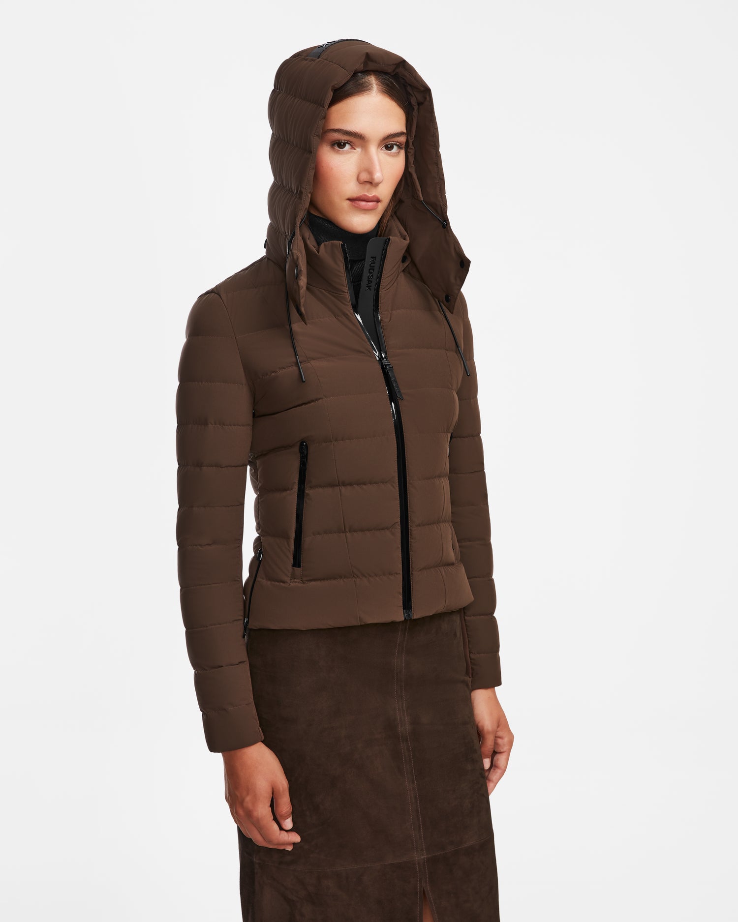 A woman wears the Rudsak MELISSA - 8124824 Espresso weatherproof down jacket with the hood up and a dark brown skirt, standing against a plain white background.