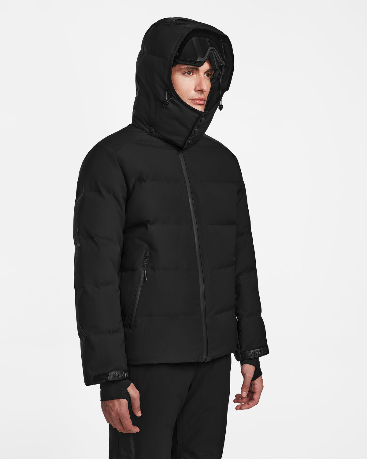 A person wearing the Rudsak MICHEL - 6124517 Black men’s ski jacket with a hood and ski goggles on their forehead stands against a plain white background.