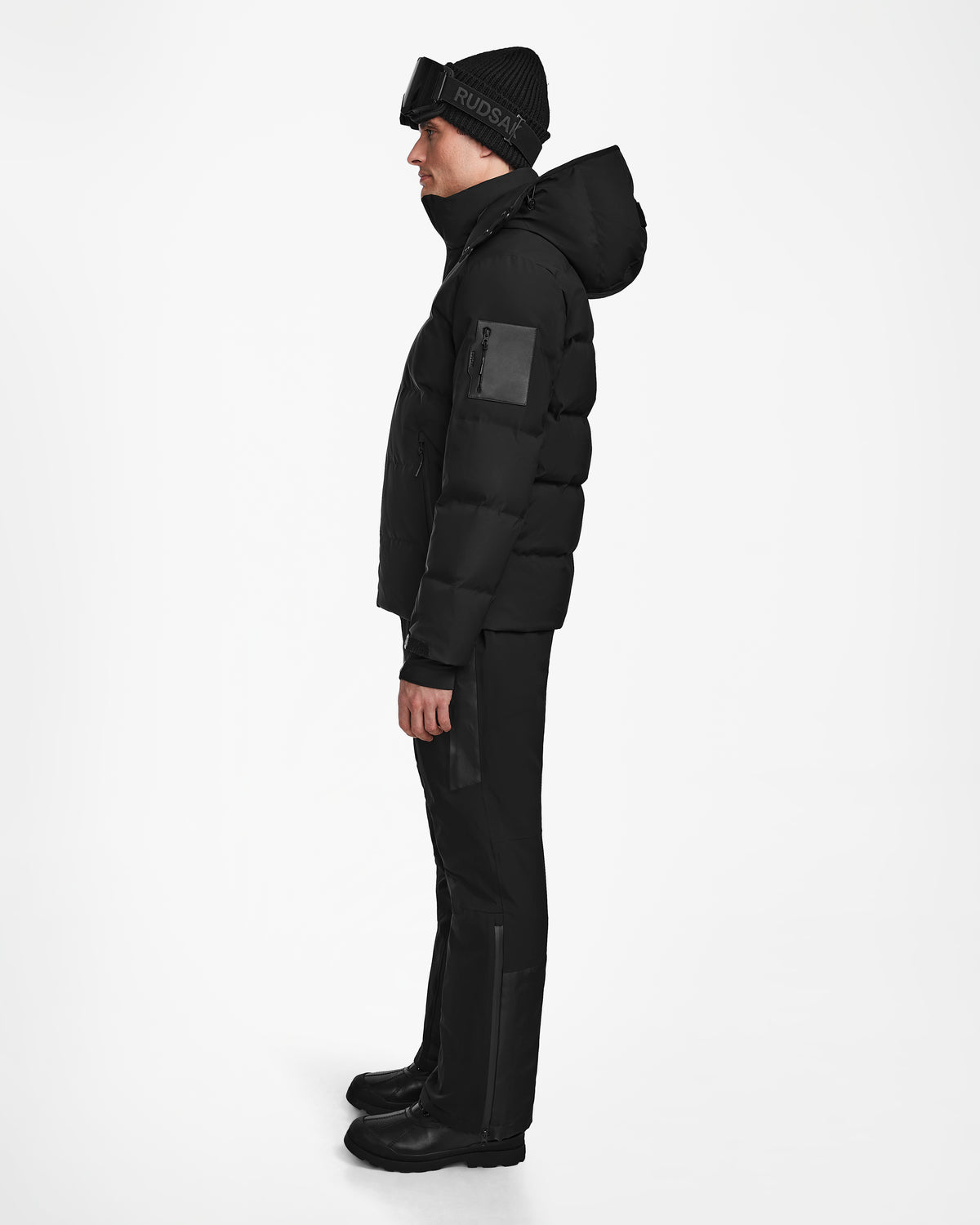 A man stands in profile wearing the Rudsak MICHEL - 6124517 Black men’s ski jacket with a hood, black pants, boots, and a beanie with ski goggles on his forehead, against a plain white background.