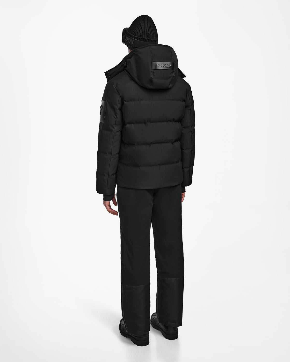 A person wearing the Rudsak MICHEL - 6124517 Black down ski jacket with RECCO rescue technology, paired with black pants, beanie, and boots, stands facing away against a plain white background.