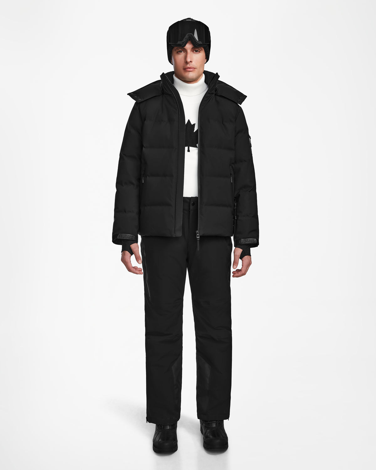 A person stands facing forward wearing the Rudsak MICHEL - 6124517 Black men’s ski jacket with RECCO rescue technology, paired with black pants, boots, gloves, a white turtleneck, and black ski goggles on their head against a plain white background.
