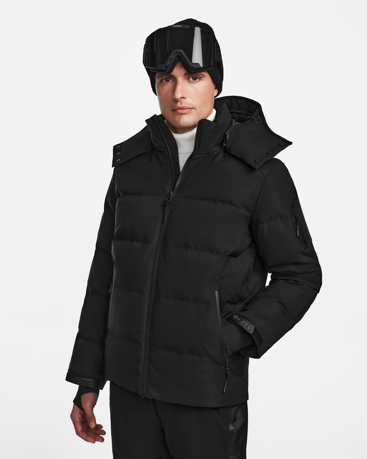 Wearing the Rudsak MICHEL - 6124517 Black down ski jacket, a person in black ski pants, a white turtleneck, black beanie, and black goggles stands against a plain white background with one hand in their pocket.