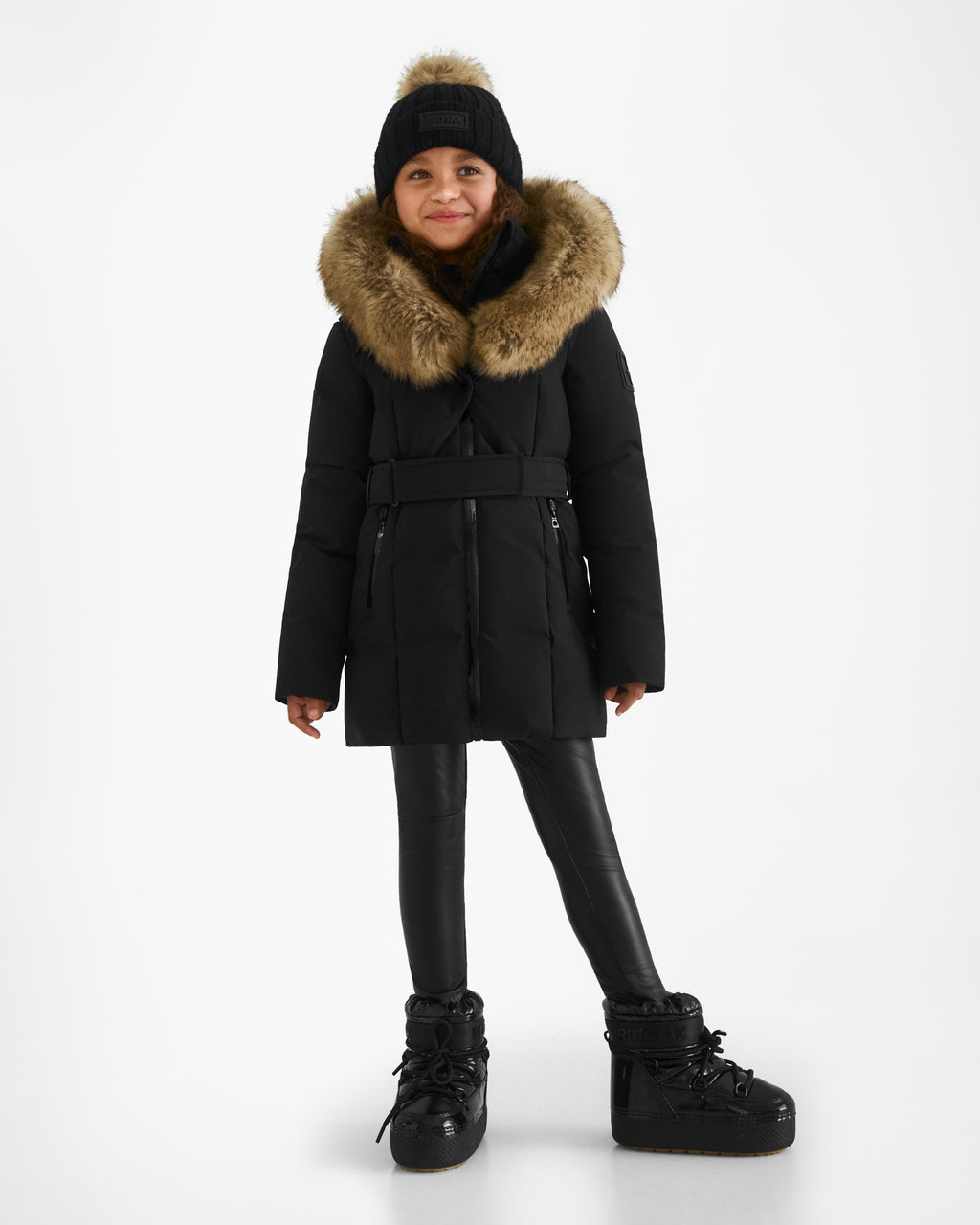 A young girl wears the Rudsak MODA K - 0124505 Black recycled nylon winter coat with a fur-lined hood, black knit pom-pom hat, leggings, and boots as she smiles against a plain white background.