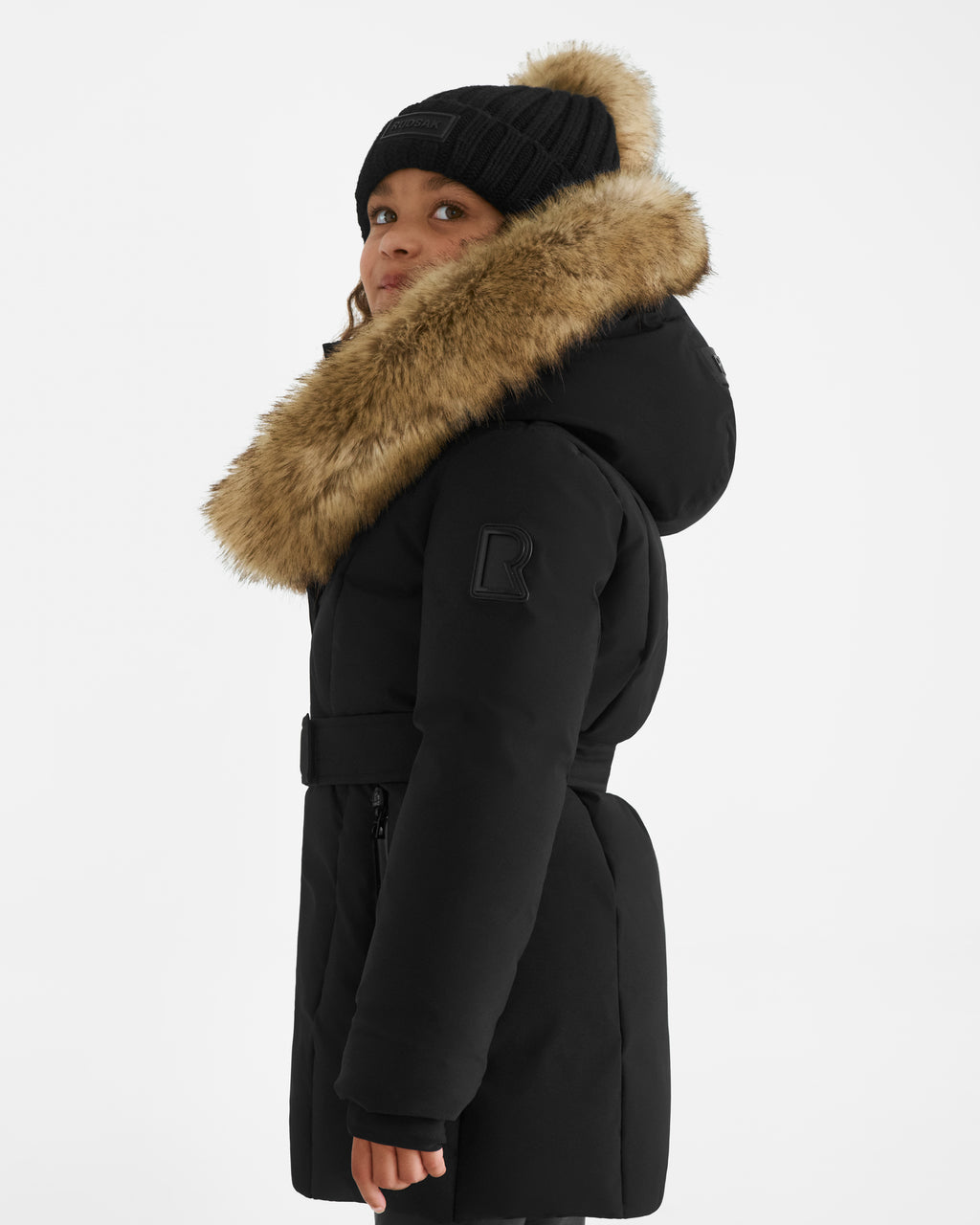 A girl in the Rudsak MODA K - 0124505 Black down parka, made from recycled nylon with a fur-lined hood, knit hat, belted waist, and R patch on the sleeve, gazes upward against a plain white background.