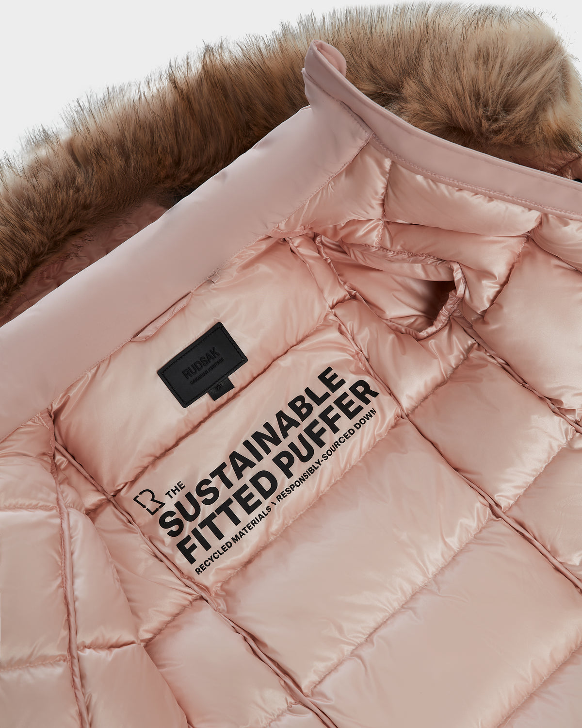 A close-up of the Rudsak MODA K - 0124505 Pastel Pink girls down parka with a brown faux fur hood. The inner lining displays bold text, The Sustainable Fitted Puffer, and highlights its recycled nylon for cozy winter wear.