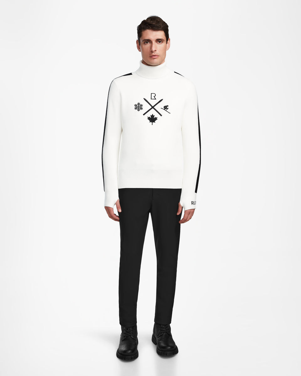A man stands against a white background wearing the Rudsak OLIVIER - 6524545 Optic White mens ski turtleneck sweater with black symbols, black pants, and shoes—a stylish winter wardrobe addition.