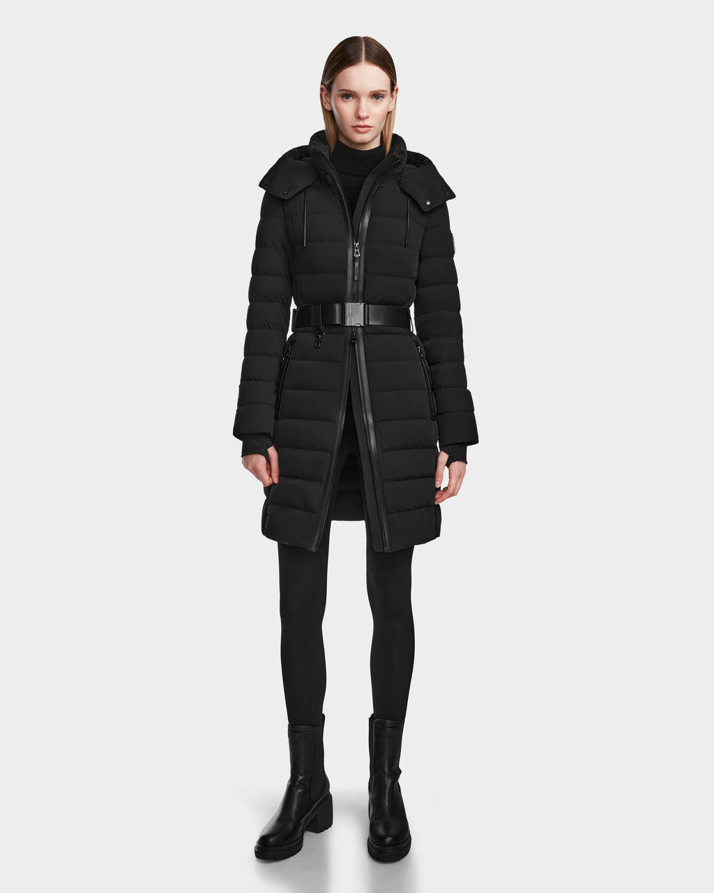 A woman faces forward in the Rudsak PELIN M - 8124814 Black belted women’s down puffer, black leggings, and ankle boots. Her straight hair is tucked behind her ears and she wears a neutral expression.