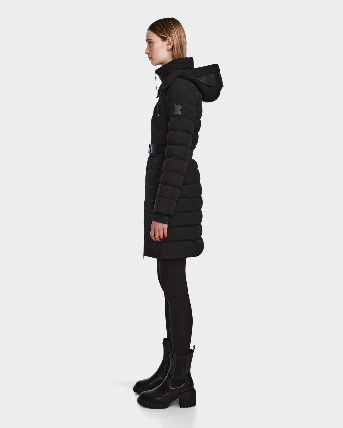 A person stands in profile against a white background, wearing the Rudsak PELIN M - 8124814 Black womens down puffer with a hood, black leggings, and black heeled boots.