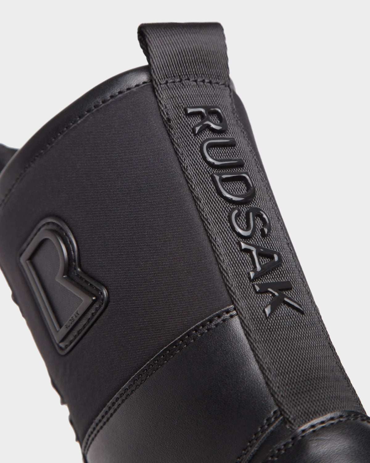 Close-up of the Rudsak RITA - L222502 Black womens winter boot with weather-treated leather, a raised RUDSAK logo on the pull tab, and an embossed R emblem on the upper, shown against a plain white background.
