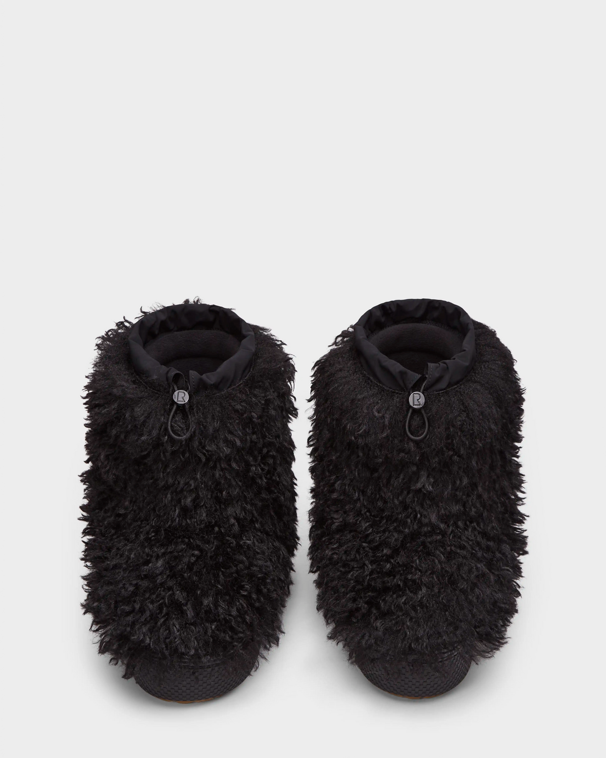 Displayed on a plain white background, the Rudsak ROSSY - 8224518 Black slippers feature black Mongolian fur, textured soles, and adjustable drawstring closures, inspired by women’s slip-on winter boots.