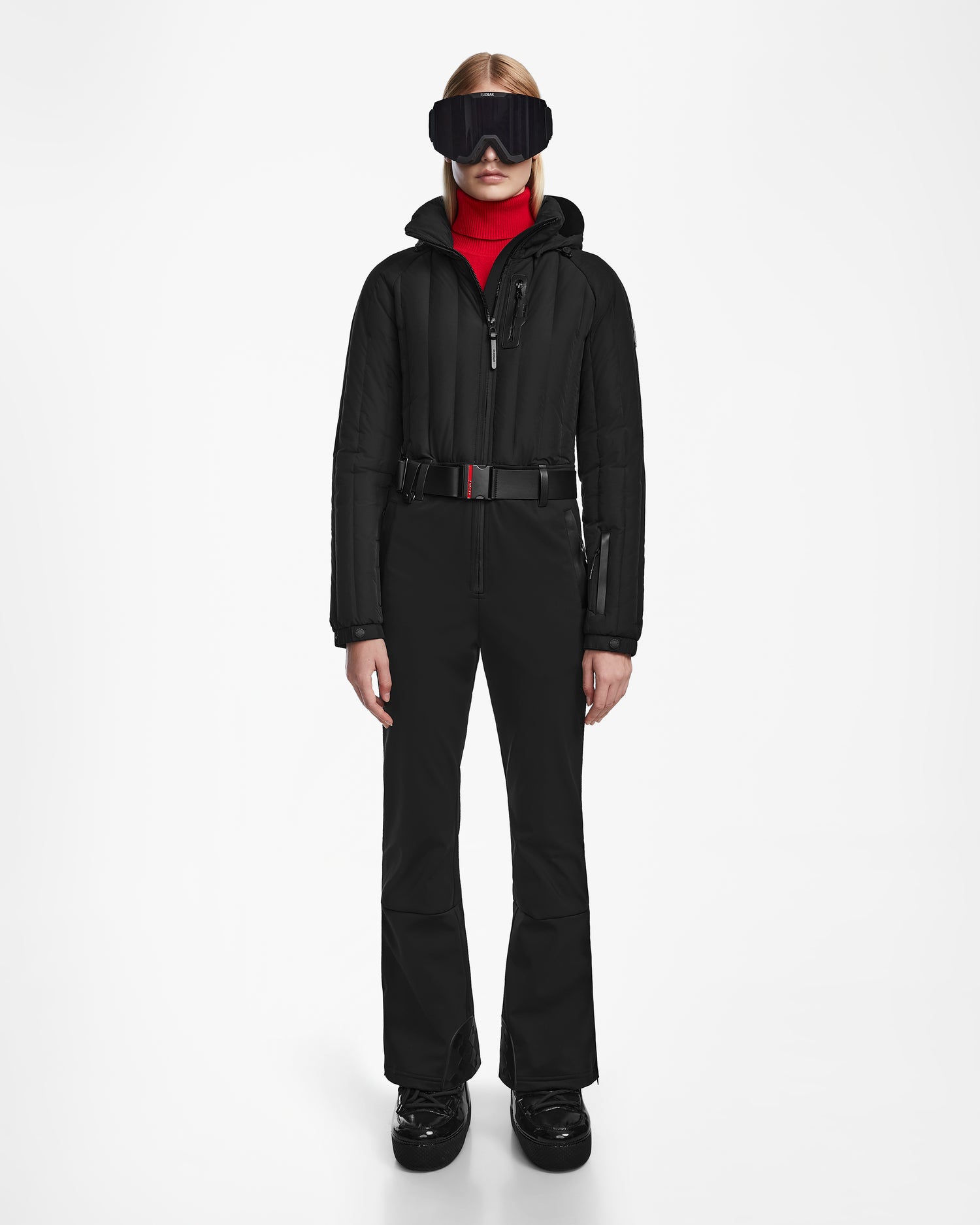 A person faces forward in the Rudsak SAGE - 8124782 Black fitted women’s ski suit with a belt, black ski boots, red turtleneck, and large black goggles against a white background. The suit is equipped with RECCO rescue technology.