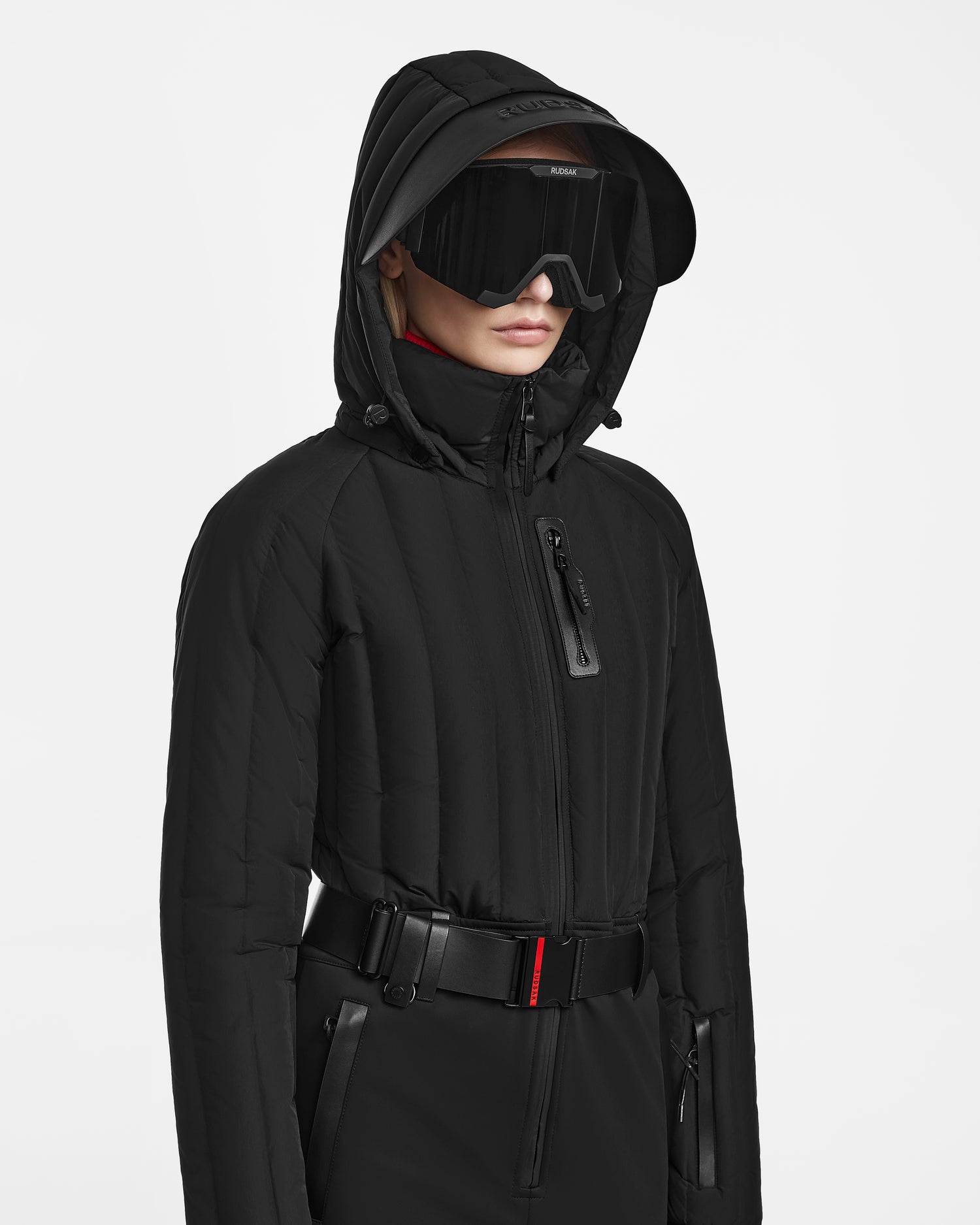 Wearing the Rudsak SAGE - 8124782 Black fitted ski suit with a black hooded jacket and large visor sunglasses, a person stands against a white background, their face partially hidden by the hood and visor.