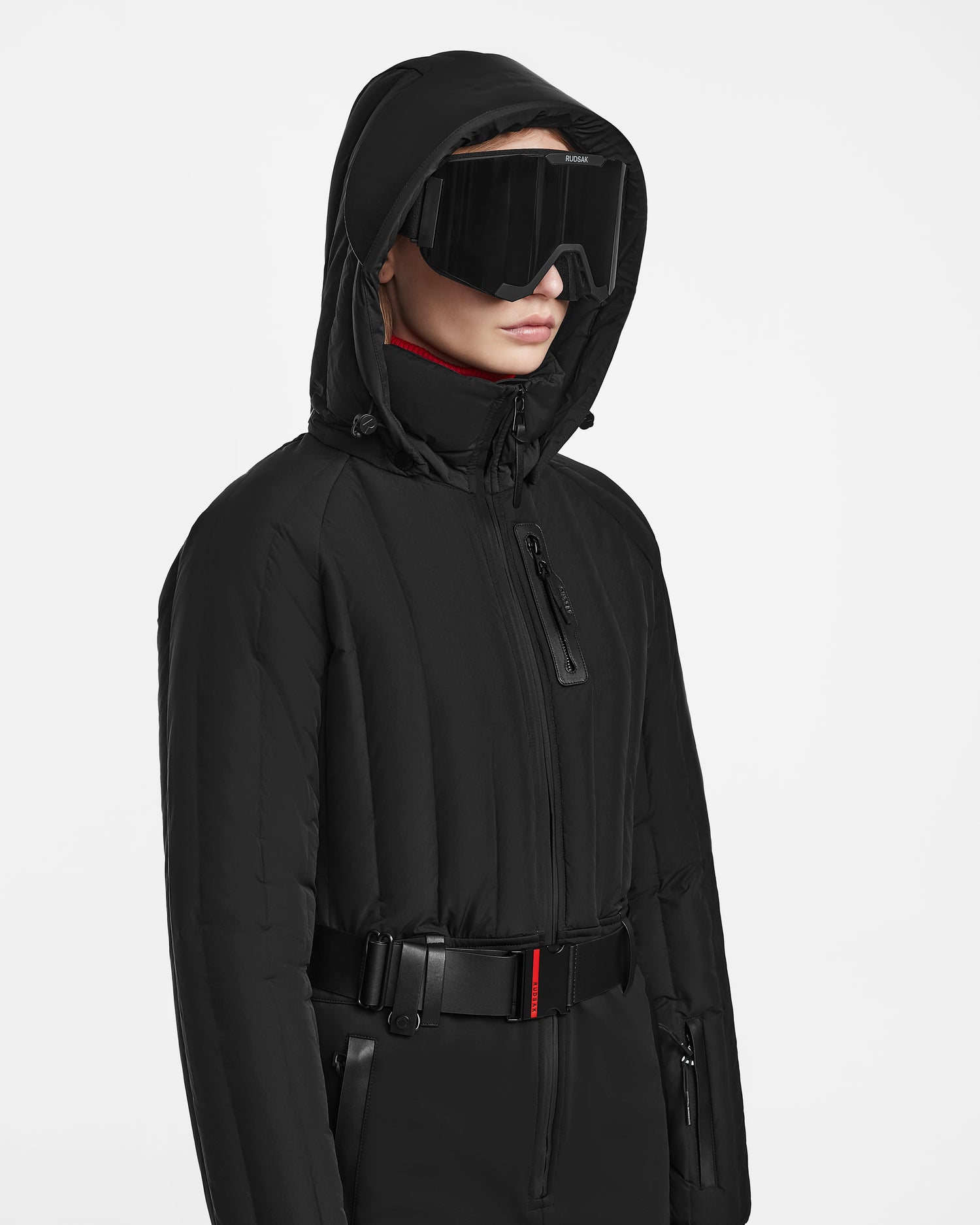 Wearing the Rudsak SAGE - 8124782 Black one-piece ski suit with a high-collared hood, black belt, and large black visor sunglasses, a person stands against a plain white background.