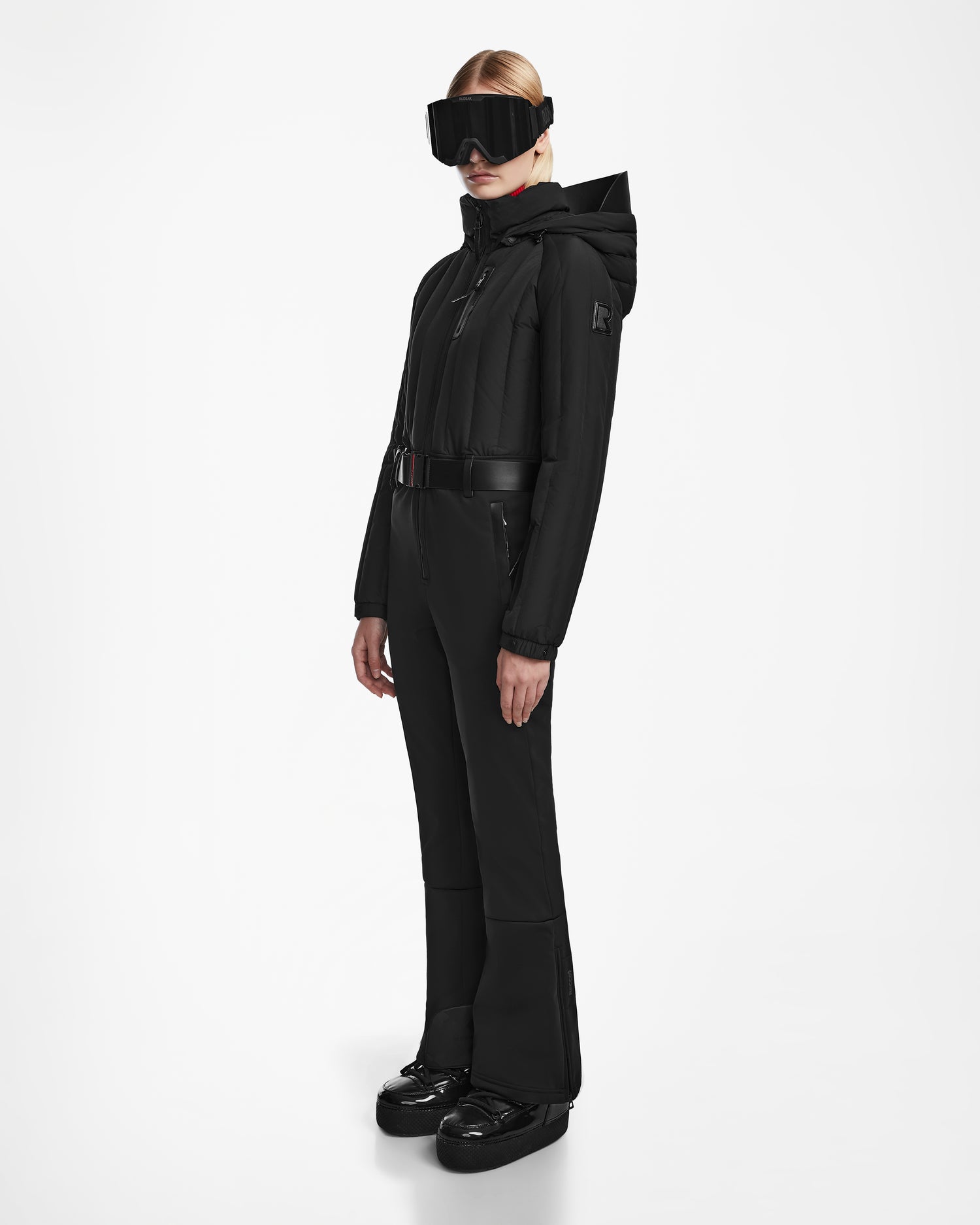 A woman models the Rudsak SAGE - 8124782 Black ski suit, featuring a belted waist, fitted one-piece design, and hood, paired with black ski goggles and shiny black boots against a plain white background.
