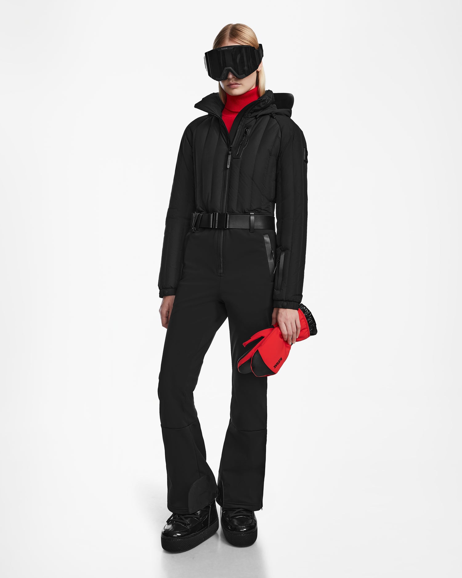 A woman stands against a white background wearing the Rudsak SAGE - 8124782 Black fitted one-piece ski suit, black boots, large black goggles, a red turtleneck, and holding red gloves.