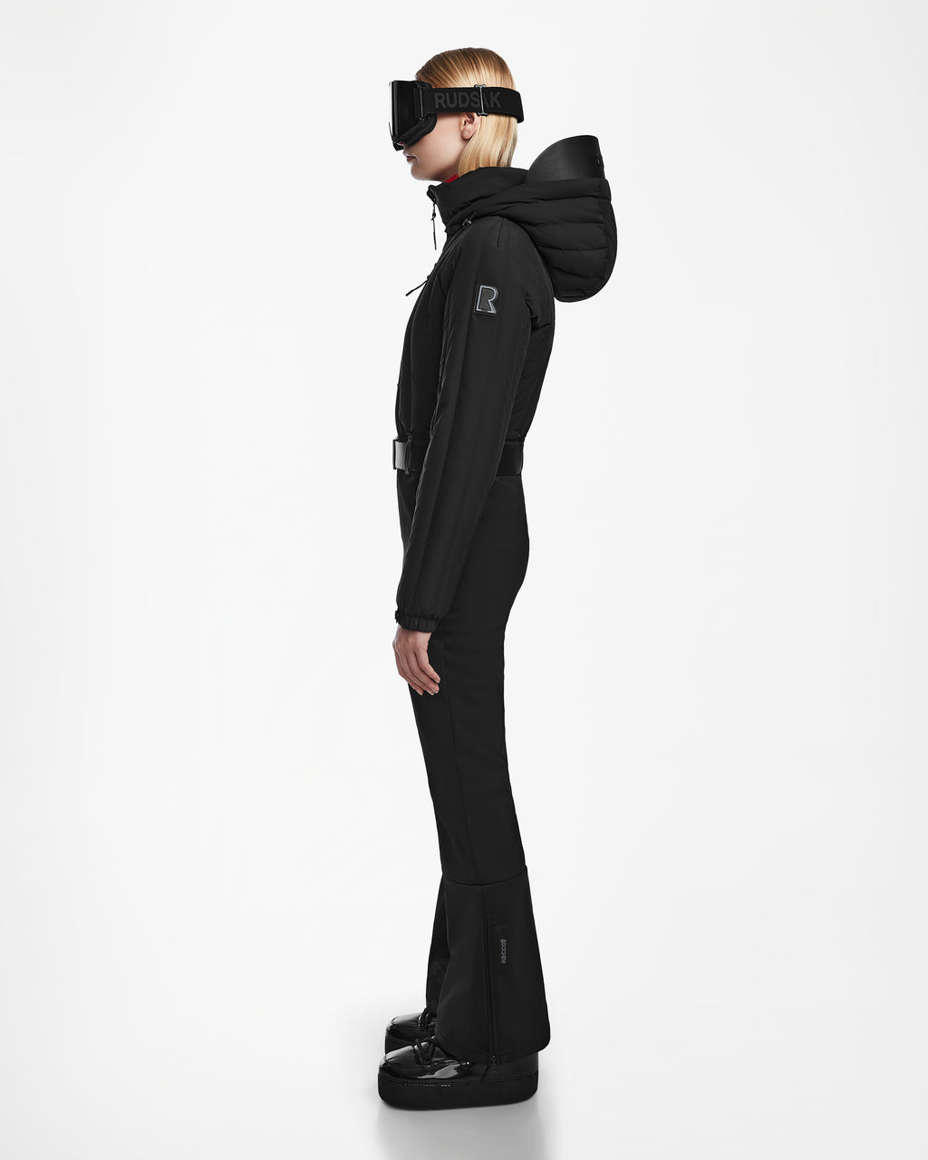 A woman in the Rudsak SAGE - 8124782 Black fitted one-piece ski suit with RECCO rescue technology, hood up, stands in profile against a white background, wearing large black ski goggles and chunky black boots.