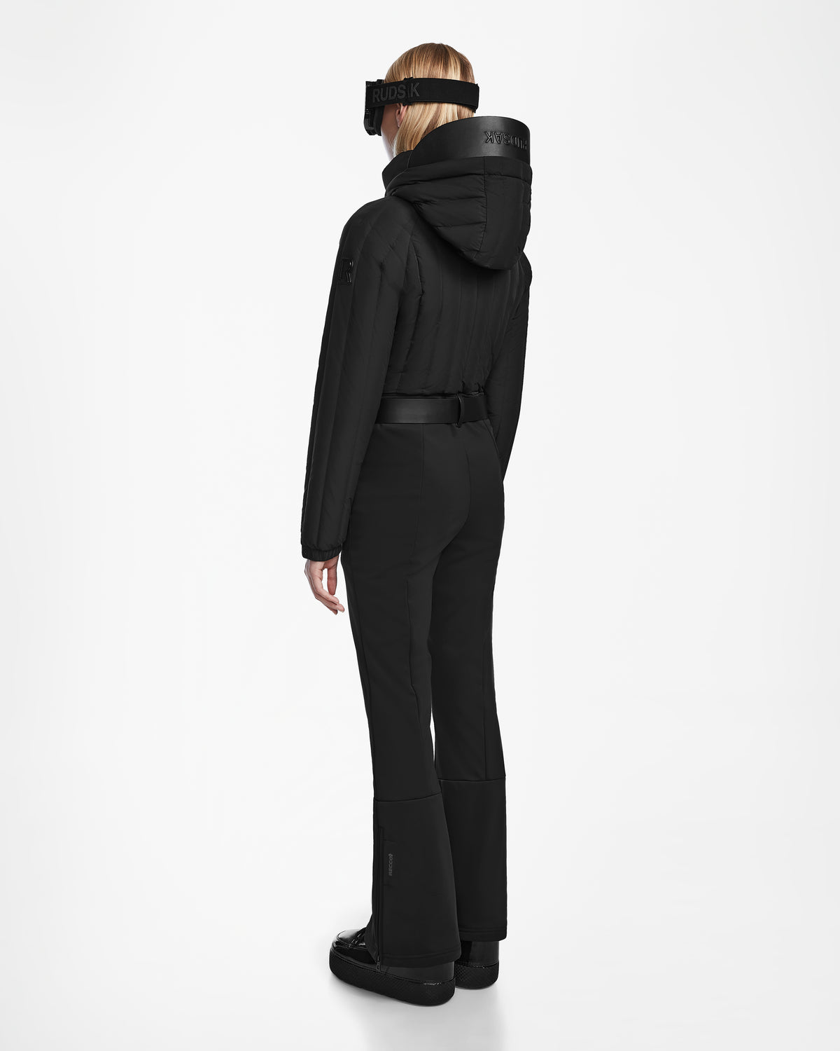 A person stands facing away in the Rudsak SAGE - 8124782 Black ski suit with RECCO rescue tech, black boots, and a black visor. The tailored, sleek outfit stands out against a plain white background.