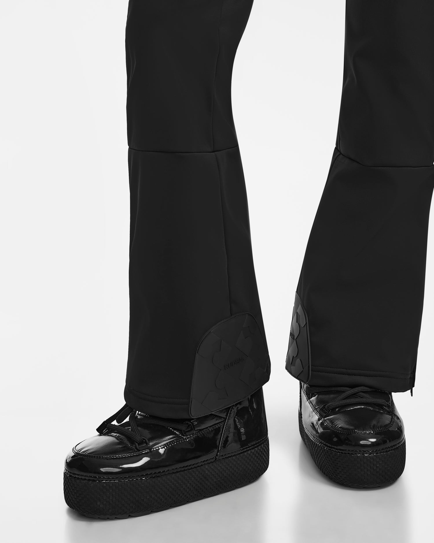 A person wears Rudsak's SAGE - 8124782 Black flared pants with ankle zippers and shiny black platform shoes, evoking a fitted ski suit style, standing on a white background.