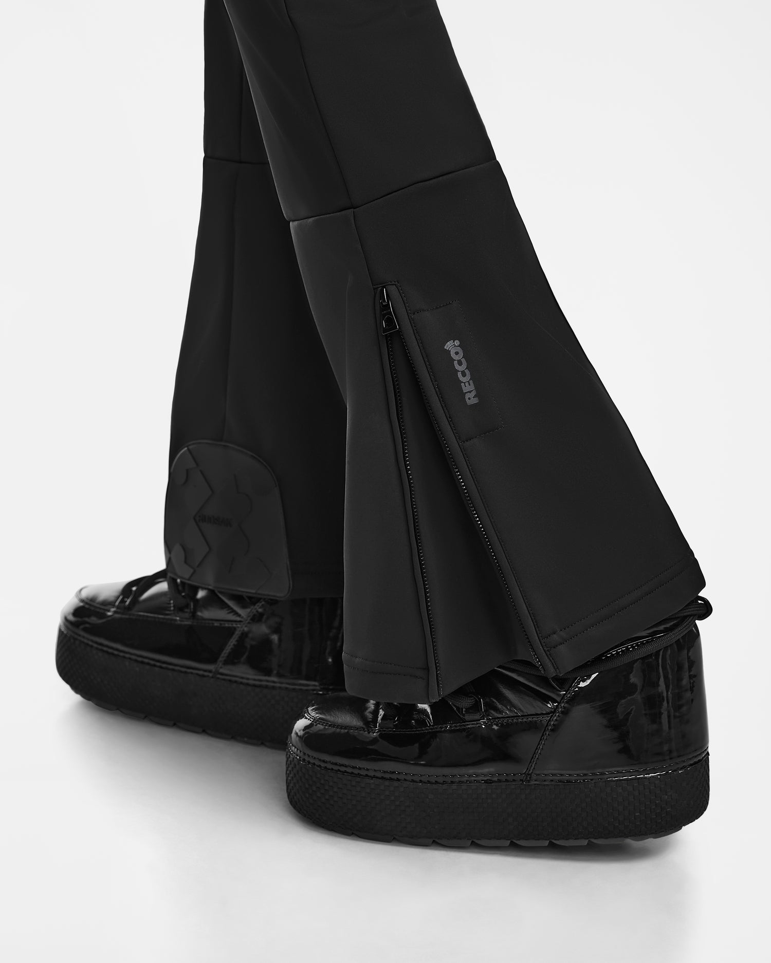 A close-up of Rudsak's SAGE - 8124782 Black flared pants with zippered openings, worn over shiny black platforms. Featuring a RECCO label and patch, these pants reflect high-performance ski suit design. Shown against a white background.