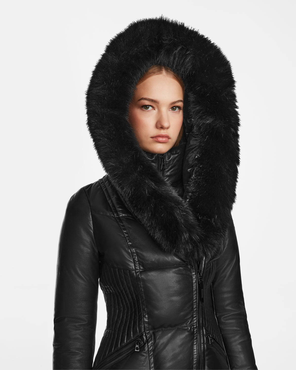 A woman in the Rudsak SHAUNA X - 8122535 Black puffer with a black leather coat and large furry hood stands facing forward against a white background, looking slightly serious.
