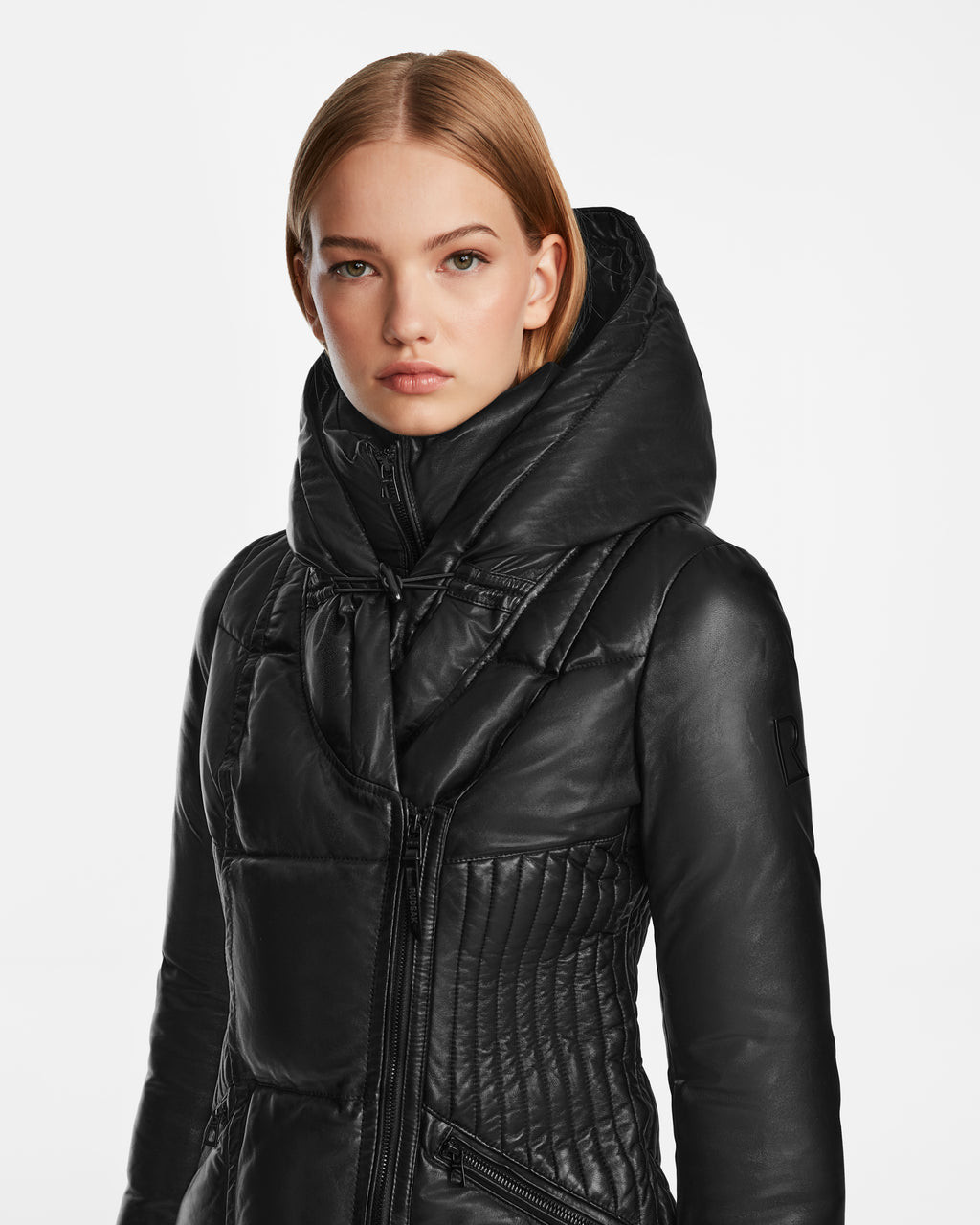 A woman with straight blonde hair wears the Rudsak SHAUNA X - 8122535 Black, a black quilted hooded lightweight leather puffer jacket. She stands against a plain white background, looking directly at the camera with a neutral expression.