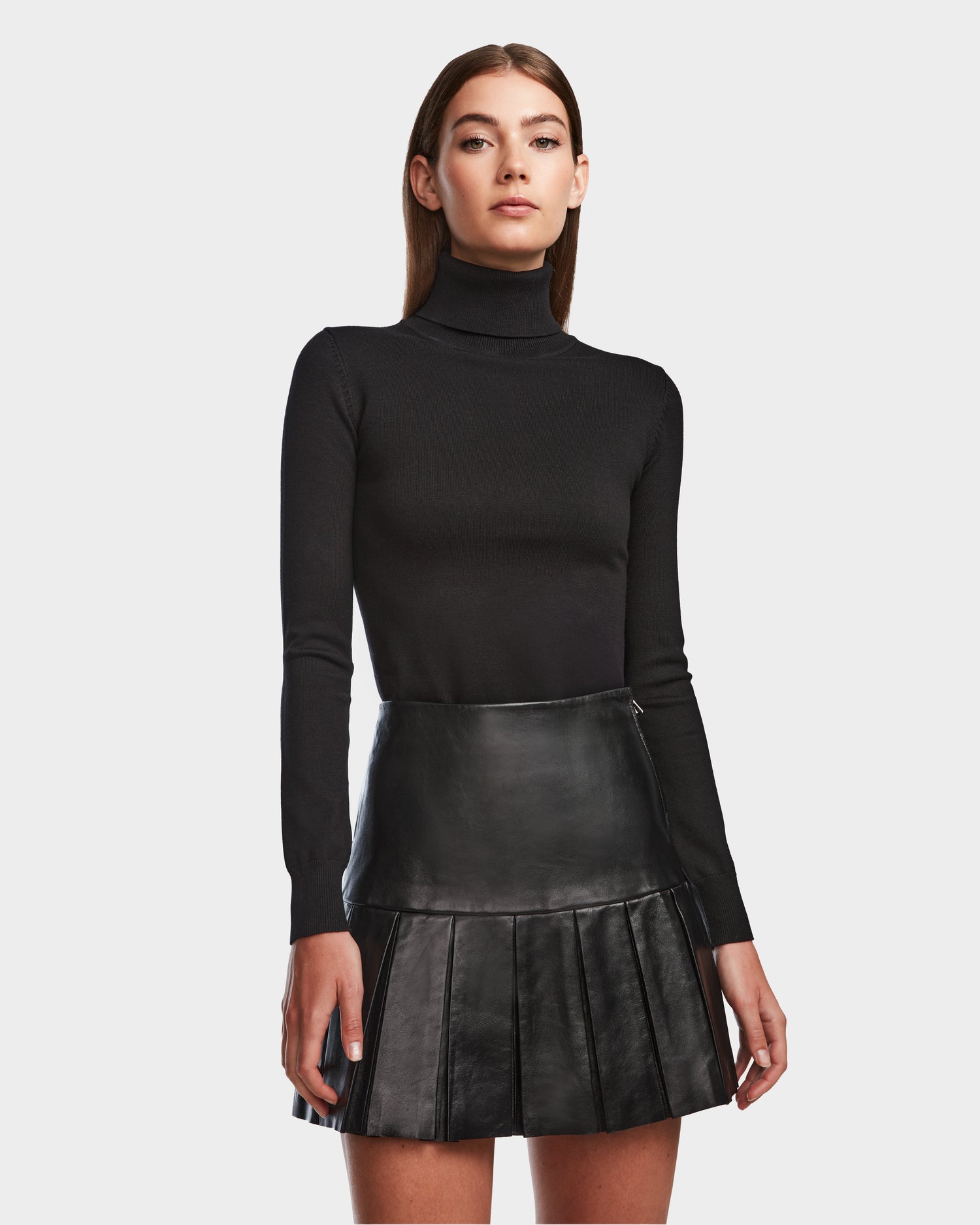 A woman with straight brown hair and a neutral expression wears a black turtleneck sweater and the Rudsak SOLANGE - 8524543 Black pleated leather skirt, standing against a plain background.
