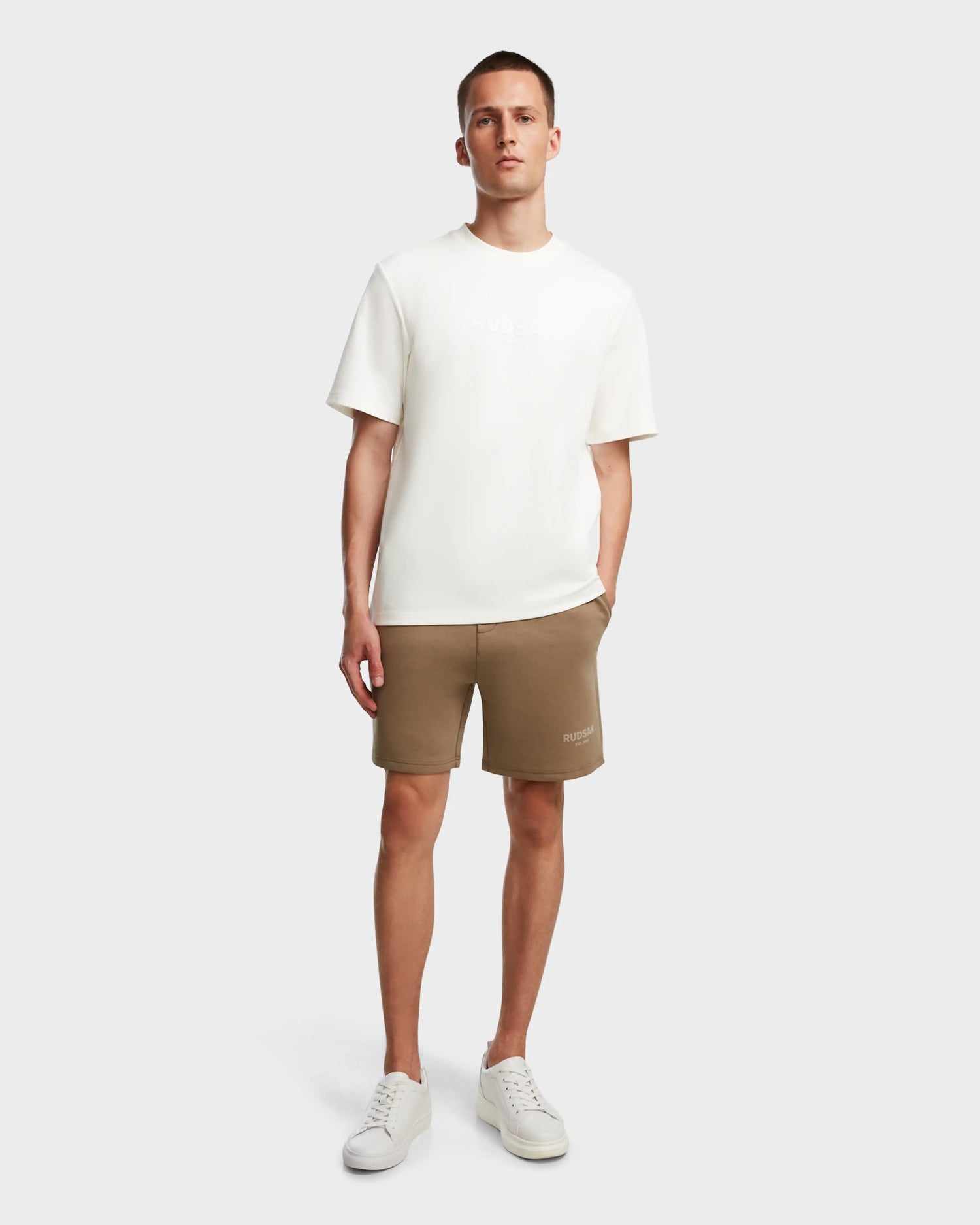 A man stands against a plain background wearing the TRAVIS - M524000 Oak brown shorts by Rudsak, paired with a white T-shirt and white sneakers. His left hand is in his pocket as he looks at the camera with a neutral expression.