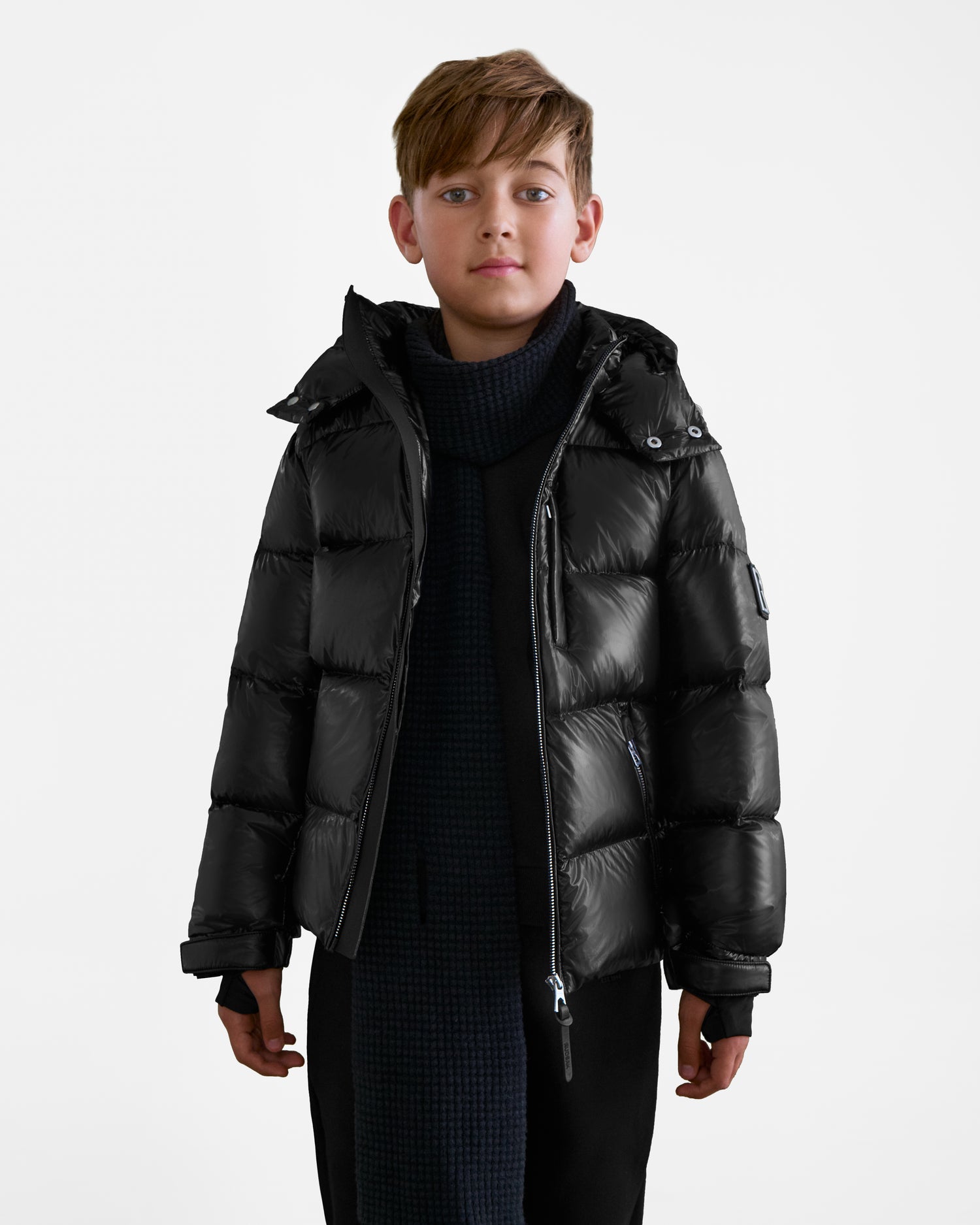 A young boy with light brown hair faces the camera, wearing a Rudsak VINCE KS - 0124502 Black unisex winter jacket and a dark textured scarf against a plain white background.