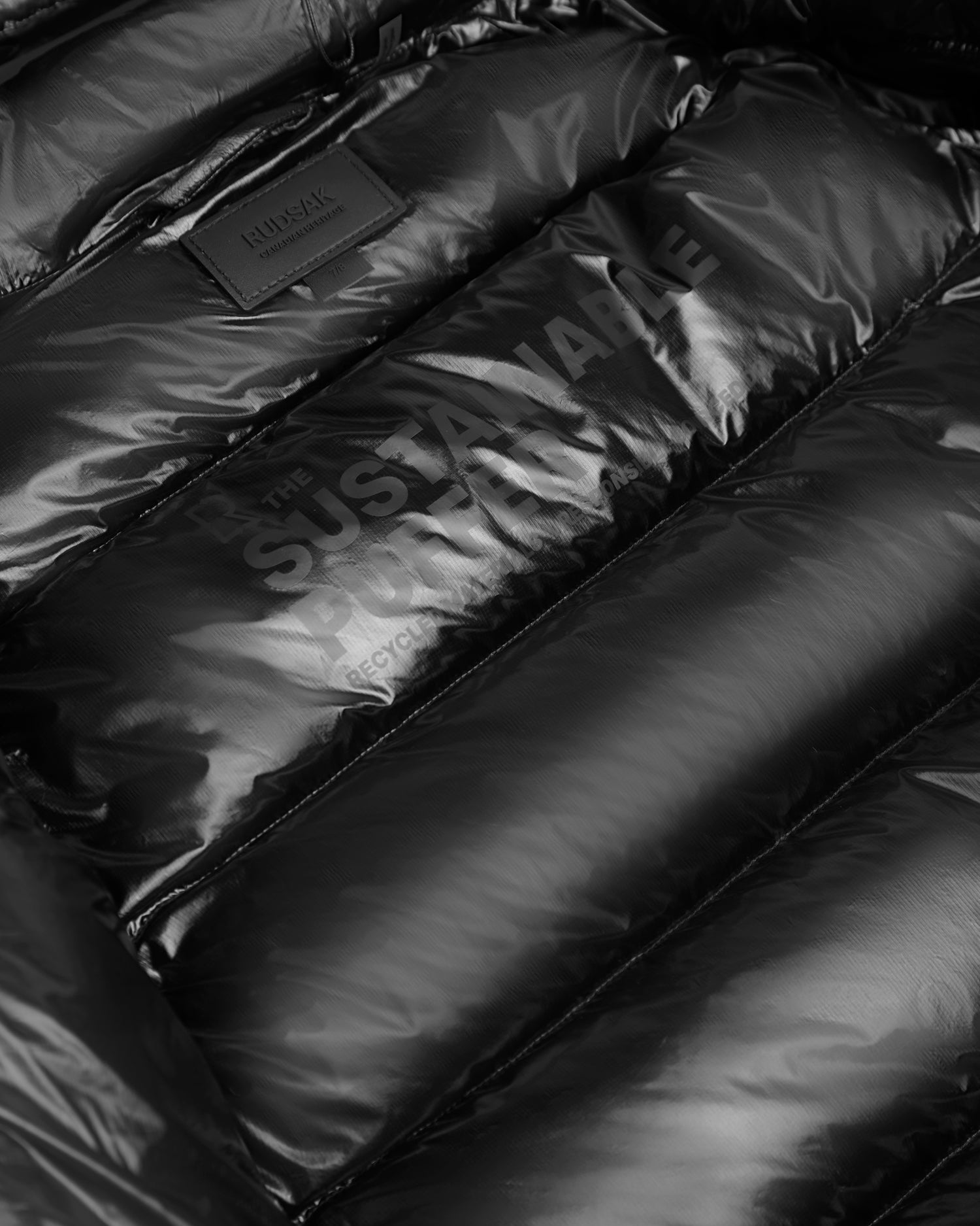 Close-up of the shiny black VINCE KS - 0124502 winter jacket by Rudsak, featuring "THE SUSTAINABLE PUFFER" and "RECYCLED" printed on the inner lining, plus a stitched brand label at the top.