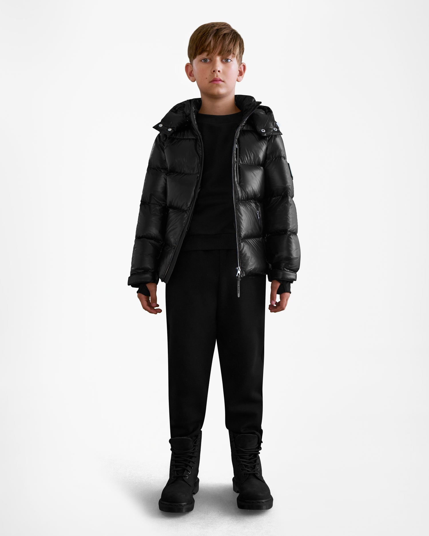 A young boy faces forward, wearing the Rudsak VINCE KS - 0124502 Black unisex winter jacket with RDS-certified down, paired with black pants and boots against a plain white background.