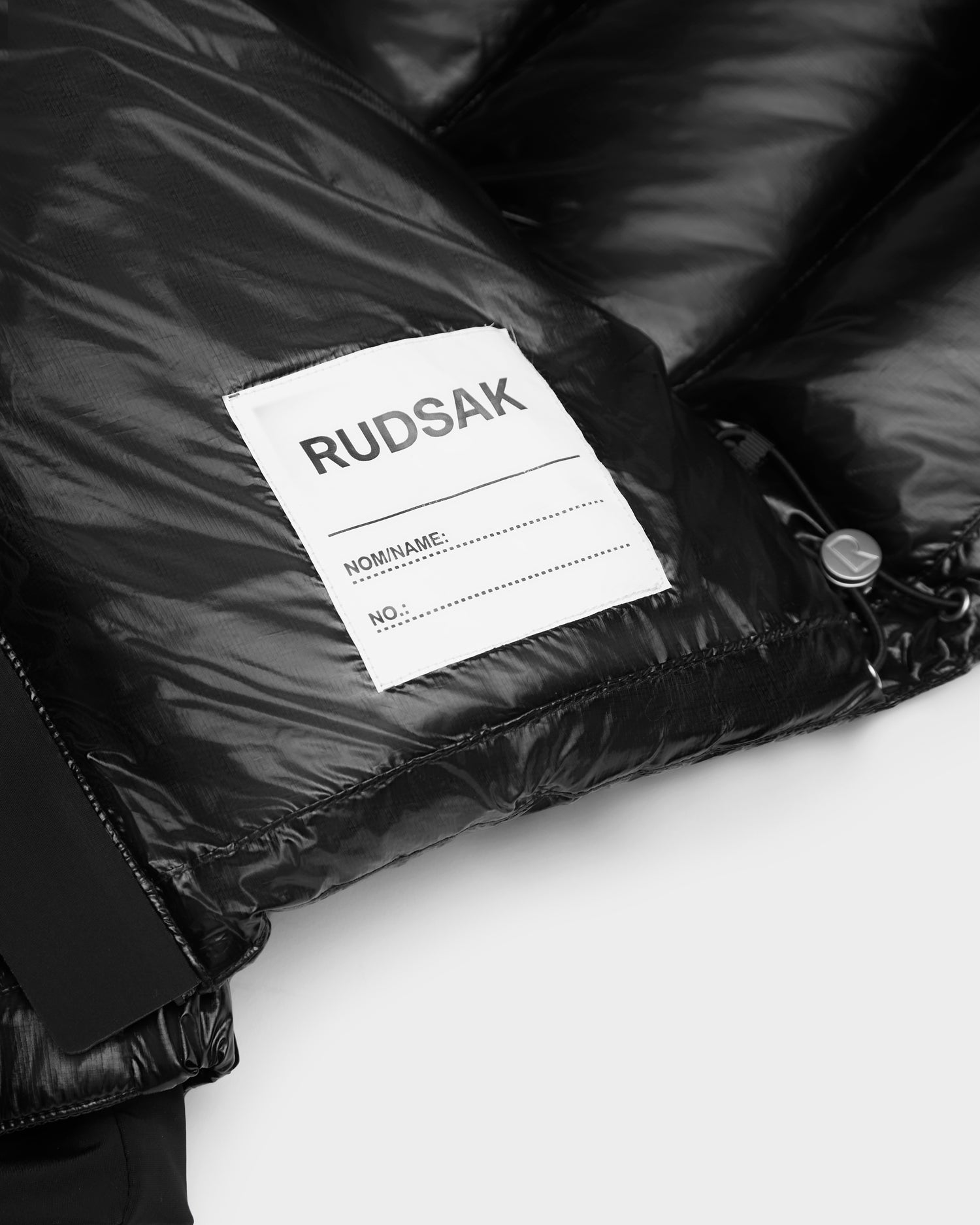 Close-up of the shiny black VINCE KS - 0124502 winter jacket by Rudsak, featuring a white RUDSAK label inside. The glossy, quilted fabric is filled with premium RDS-certified down for exceptional style and warmth.