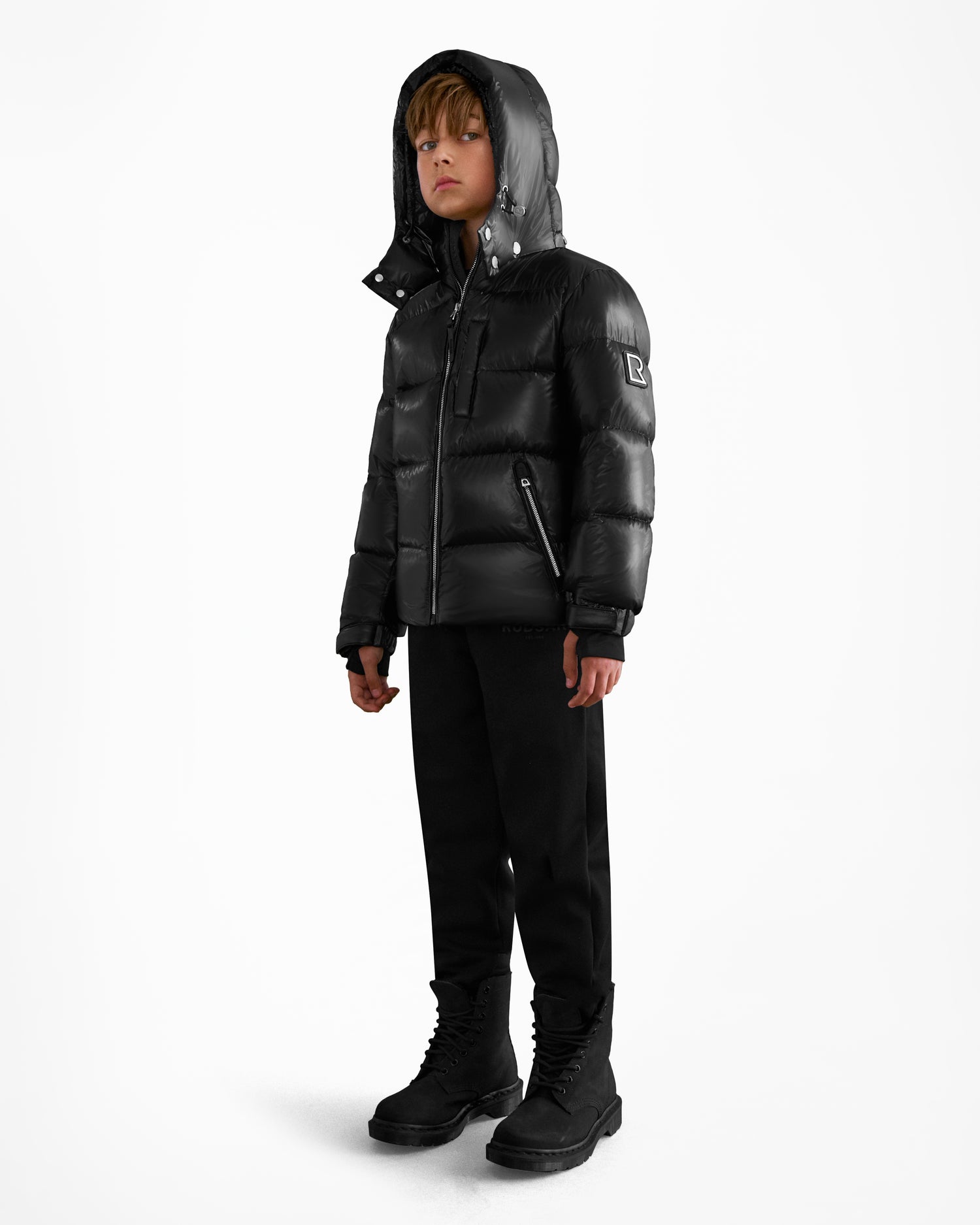 A young boy, wearing the Rudsak VINCE KS - 0124502 Black unisex winter jacket with RDS-certified down, along with black pants and boots, stands slightly turned to the side against a plain white background.