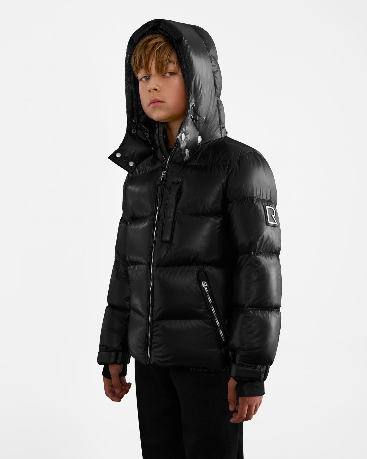 A young boy wears the Rudsak VINCE KS - 0124502 Black down puffer jacket with hood, standing against a plain white background and looking slightly to the side with a neutral expression.