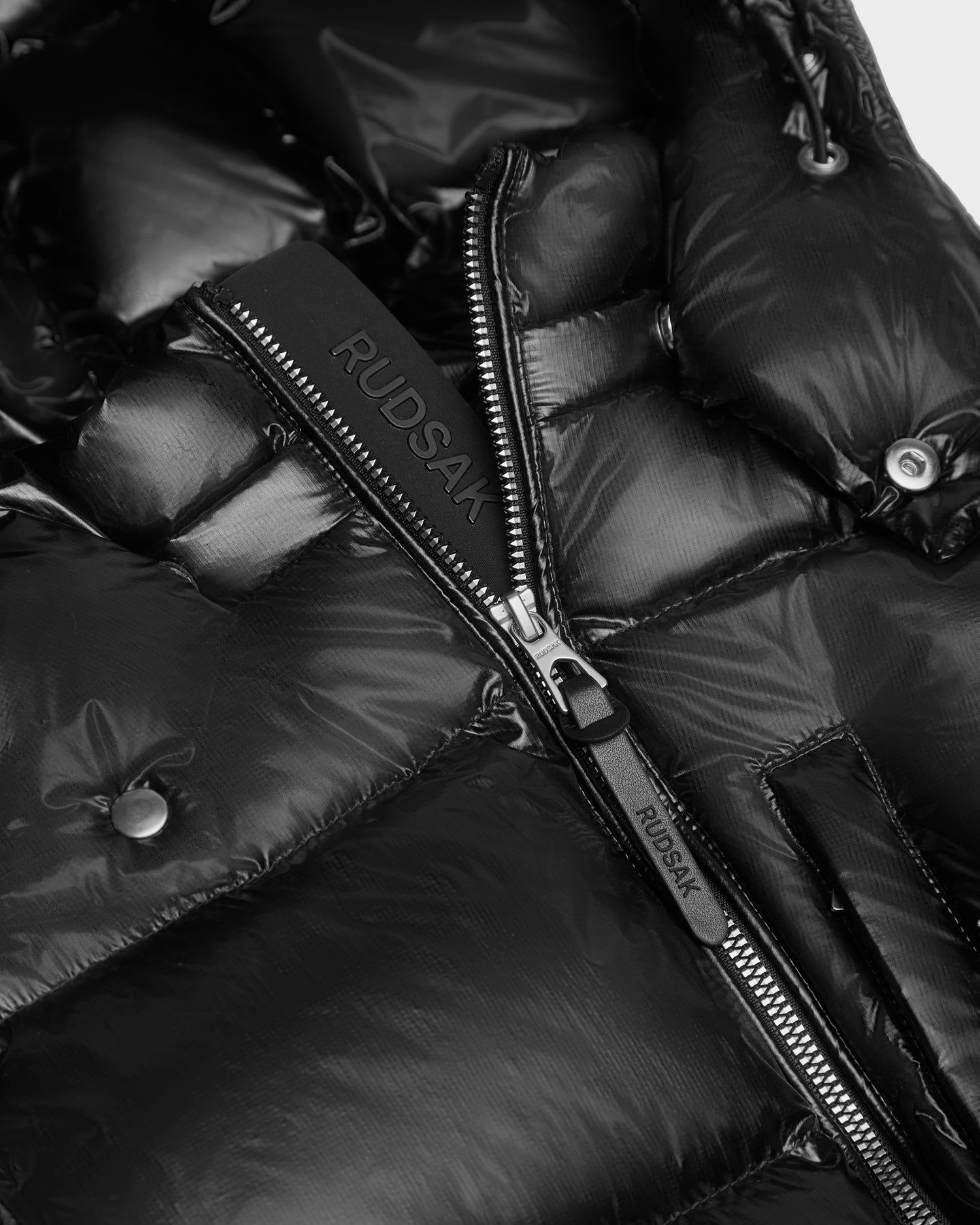 Close-up of the VINCE KS - 0124502 Black unisex winter jacket by Rudsak. Features glossy black fabric, a partially open zipper with branded pull and leather tab, snap button details, and RDS-certified down fill.