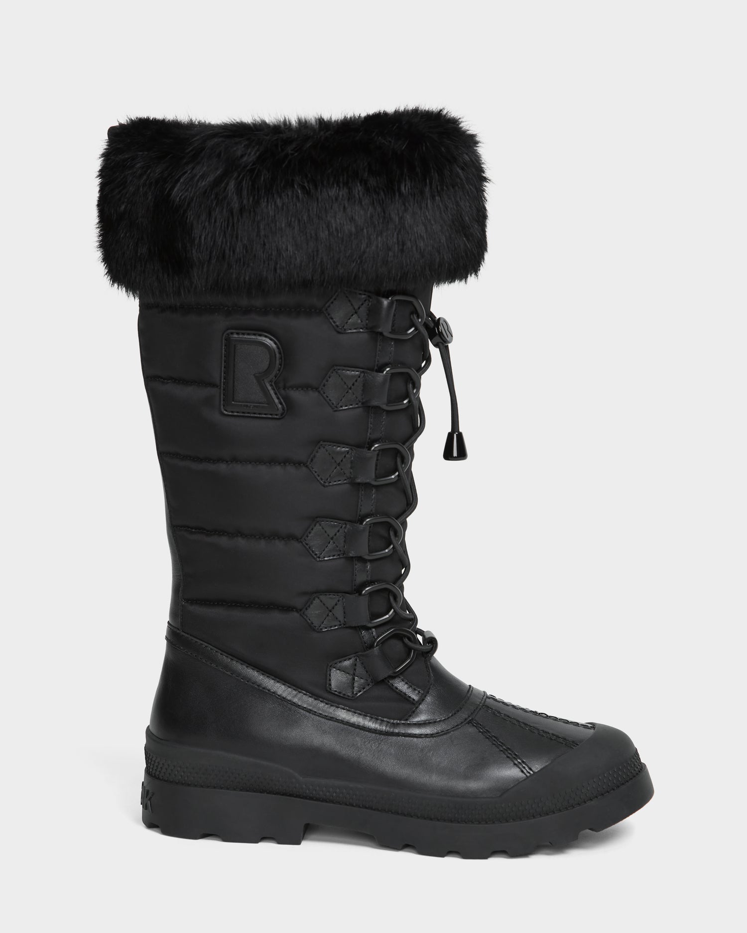 The WINDY - L224507 Black by Rudsak is a tall black women’s winter boot with faux fur trim, quilted sides, front laces, a rugged rubber sole for cold-weather comfort, and a raised R logo on the side.