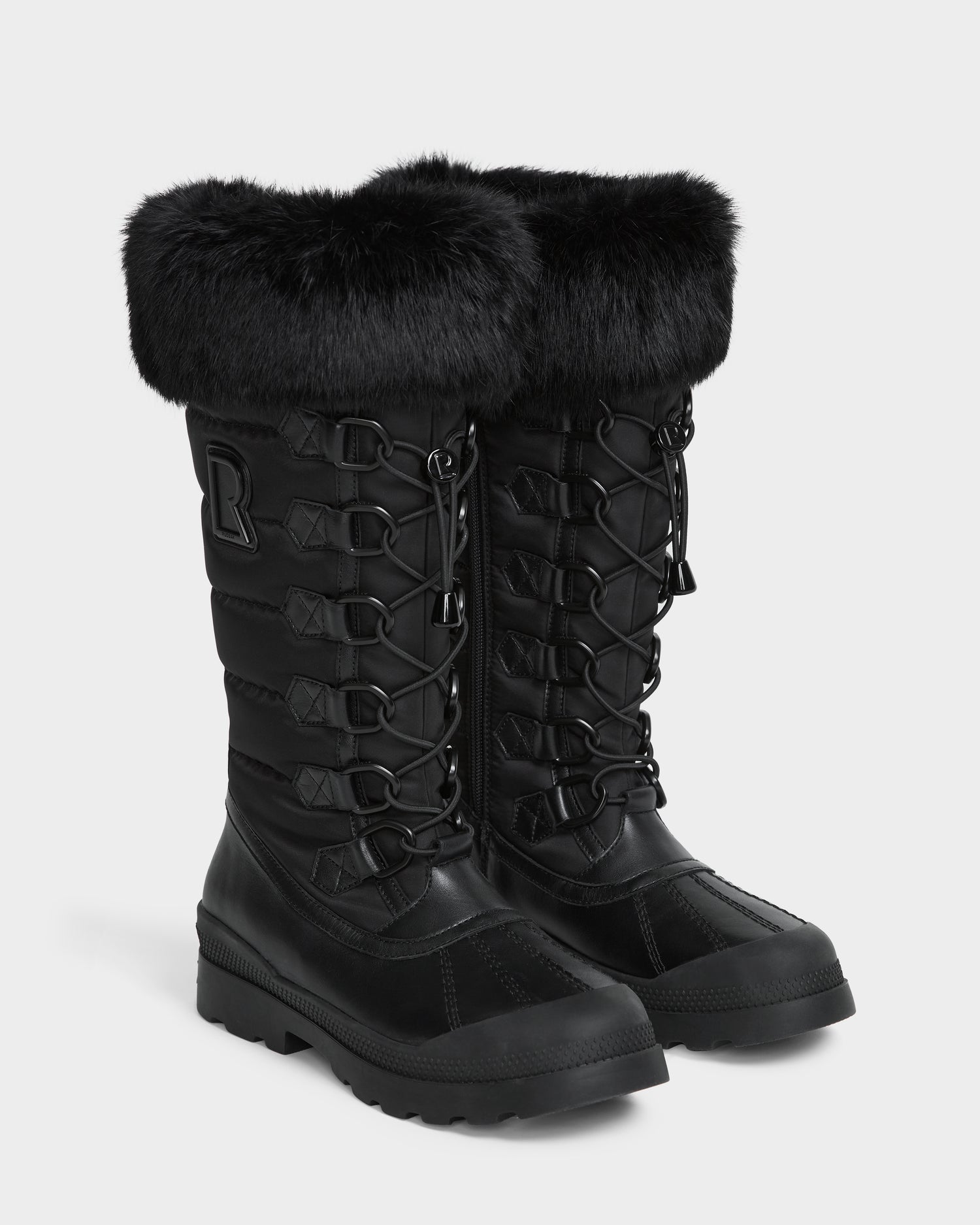 The WINDY - L224507 Black by Rudsak are tall women’s winter boots with faux fur trim, quilted design, lace-up fronts, rugged soles for warmth, and an “R” stitched on the side near the top.