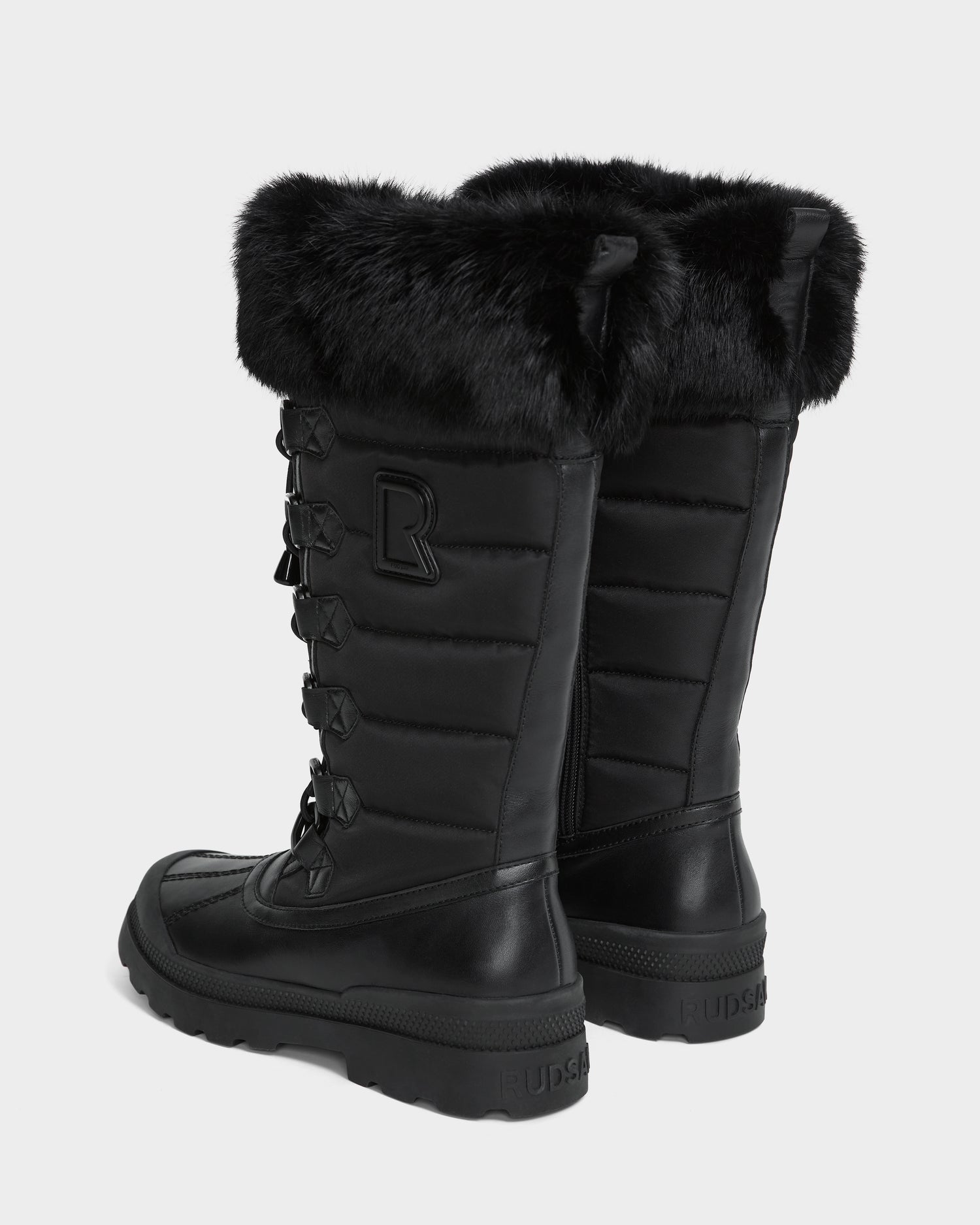 The Rudsak WINDY - L224507 Black women’s boots feature a tall design with faux fur trim, quilted texture, lace-up fronts, and sturdy rubber soles for winter comfort. Shown against a light background.