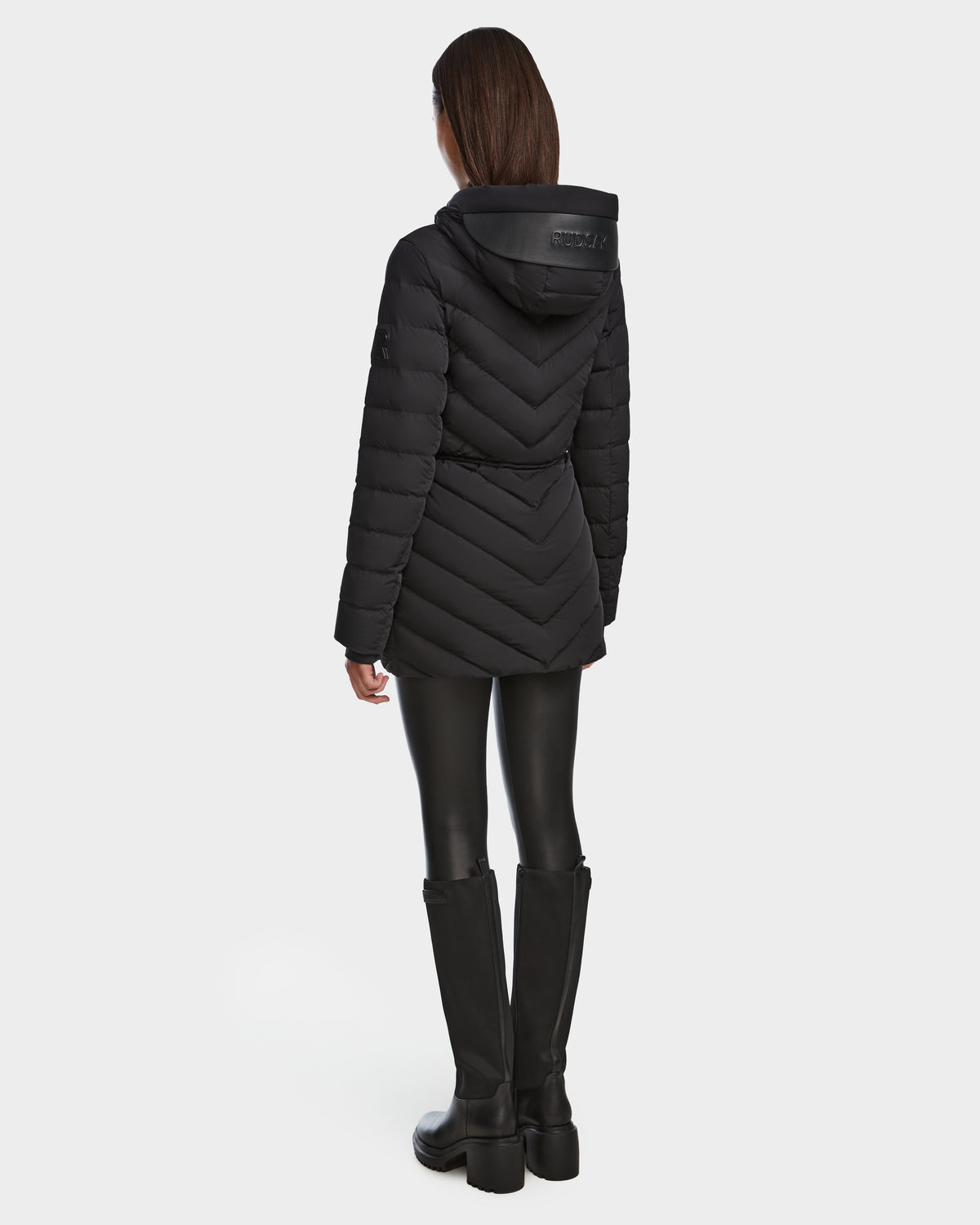 A woman stands facing away, wearing the Rudsak FRANCINE - 8124722 Black womens light down puffer jacket with chevron quilting, paired with black leggings and knee-high black boots against a plain white background.