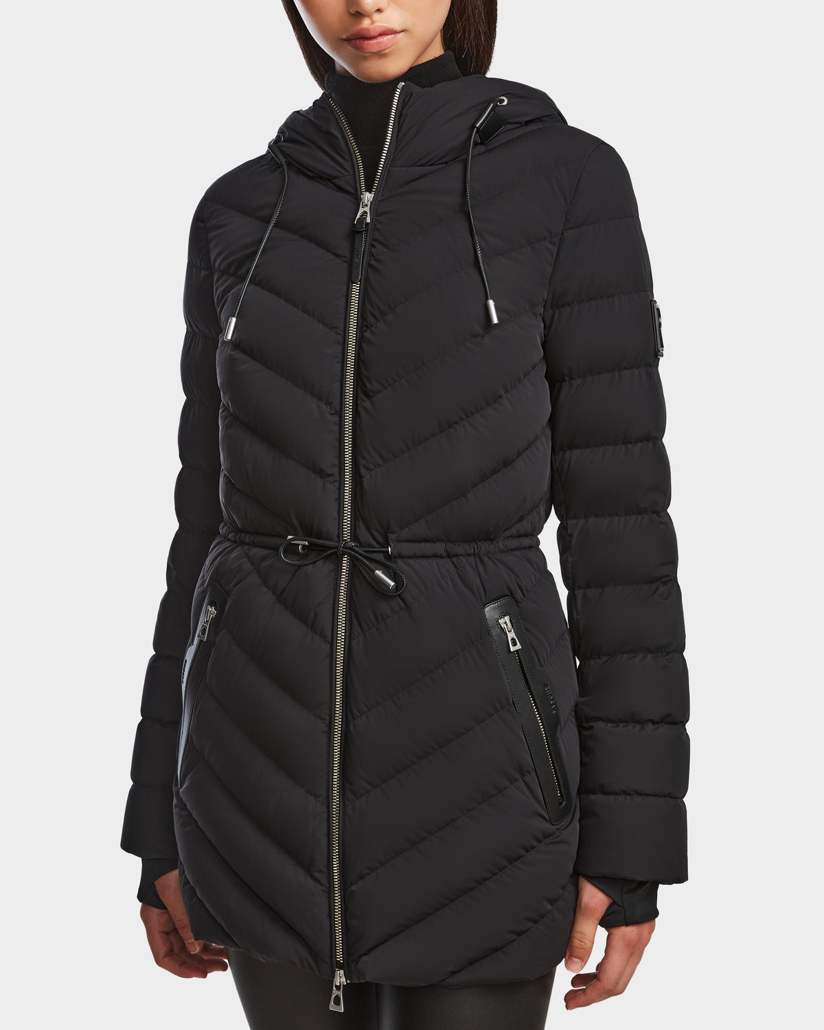 A woman wears the Rudsak FRANCINE - 8124722 Black long, hooded puffer jacket with front zipper, drawstring waist, and zippered pockets against a plain white background.
