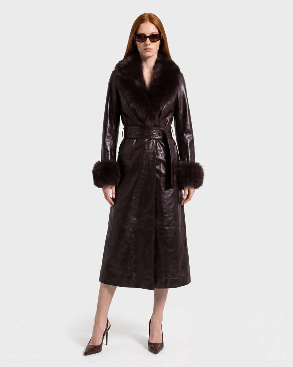 A woman stands against a plain light background, wearing the Rudsak VIVIANNE - 8126933 Espresso long, shiny dark brown leather trench featuring a luxurious faux fur collar and cuffs, paired with matching pointed heels and dark sunglasses.